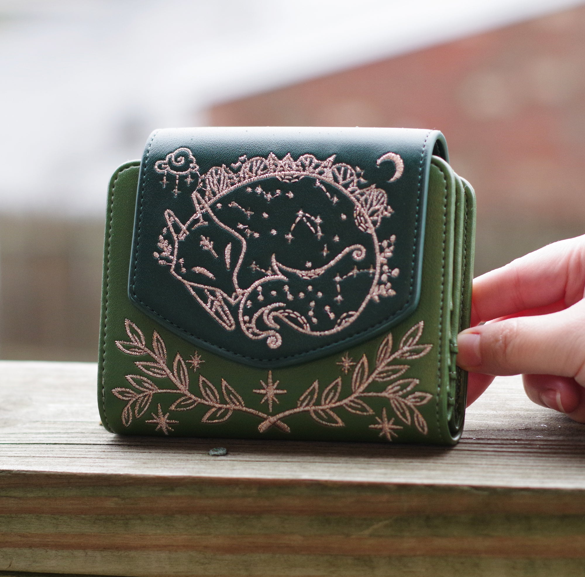 Green faux leather wallet with rose gold embroidery of a sleeping fox, floral vines, stars, and constellations. Held outdoors in natural lighting.