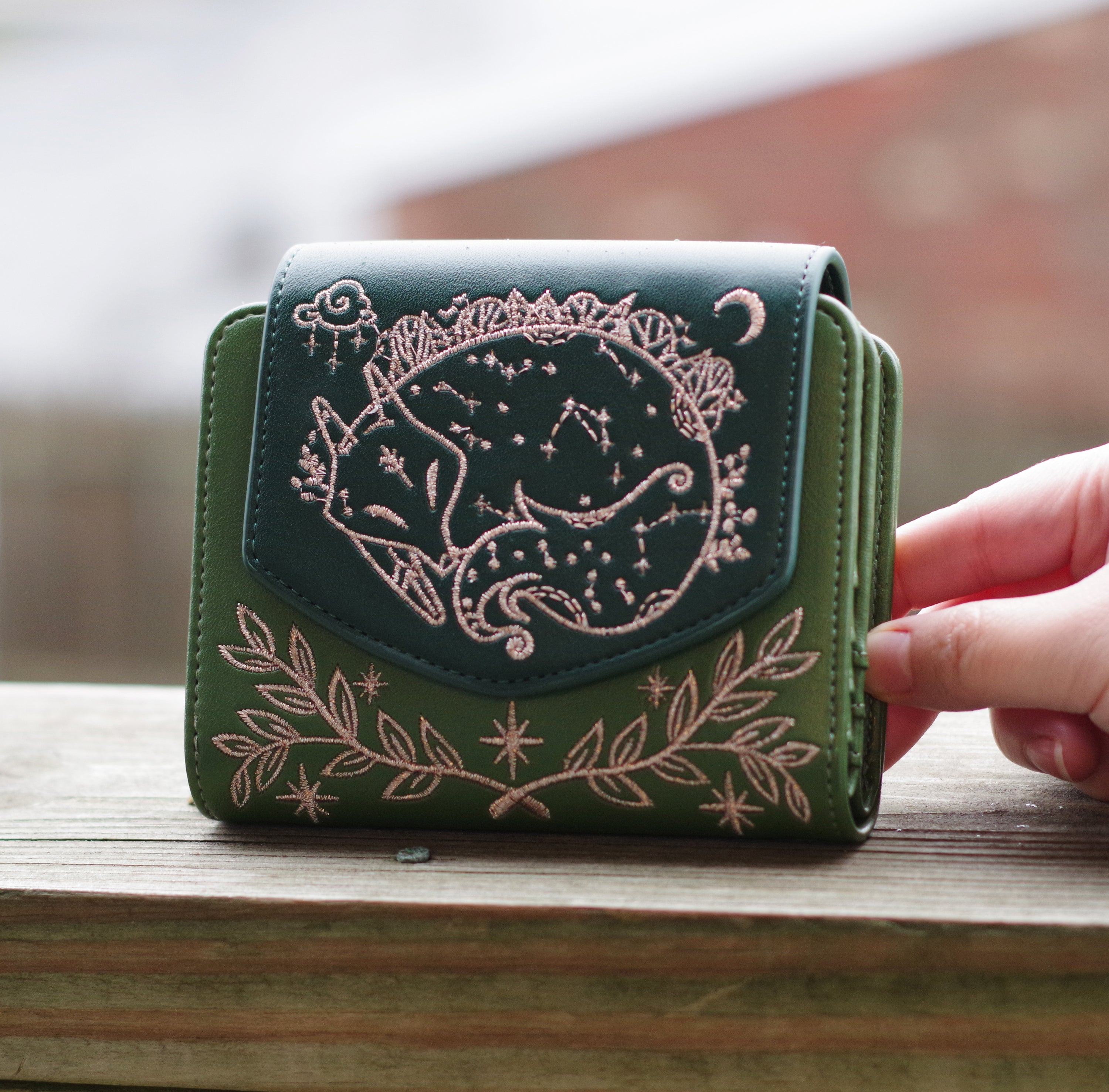 Green faux leather wallet with rose gold embroidery of a sleeping fox, floral vines, stars, and constellations. Held outdoors in natural lighting.
