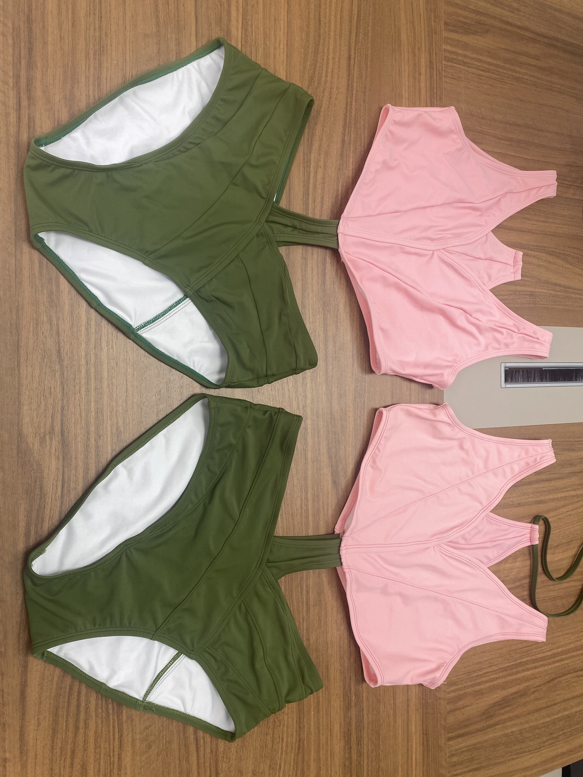 Two prototype sets of the Tulip Swimsuit laid flat on a wooden surface. Each set includes a green bottom shaped like overlapping leaves and a pink petal-shaped top. The suits are unassembled, showing separate front and back panels. The fabric has a soft matte finish, and the pieces are arranged side by side for comparison.