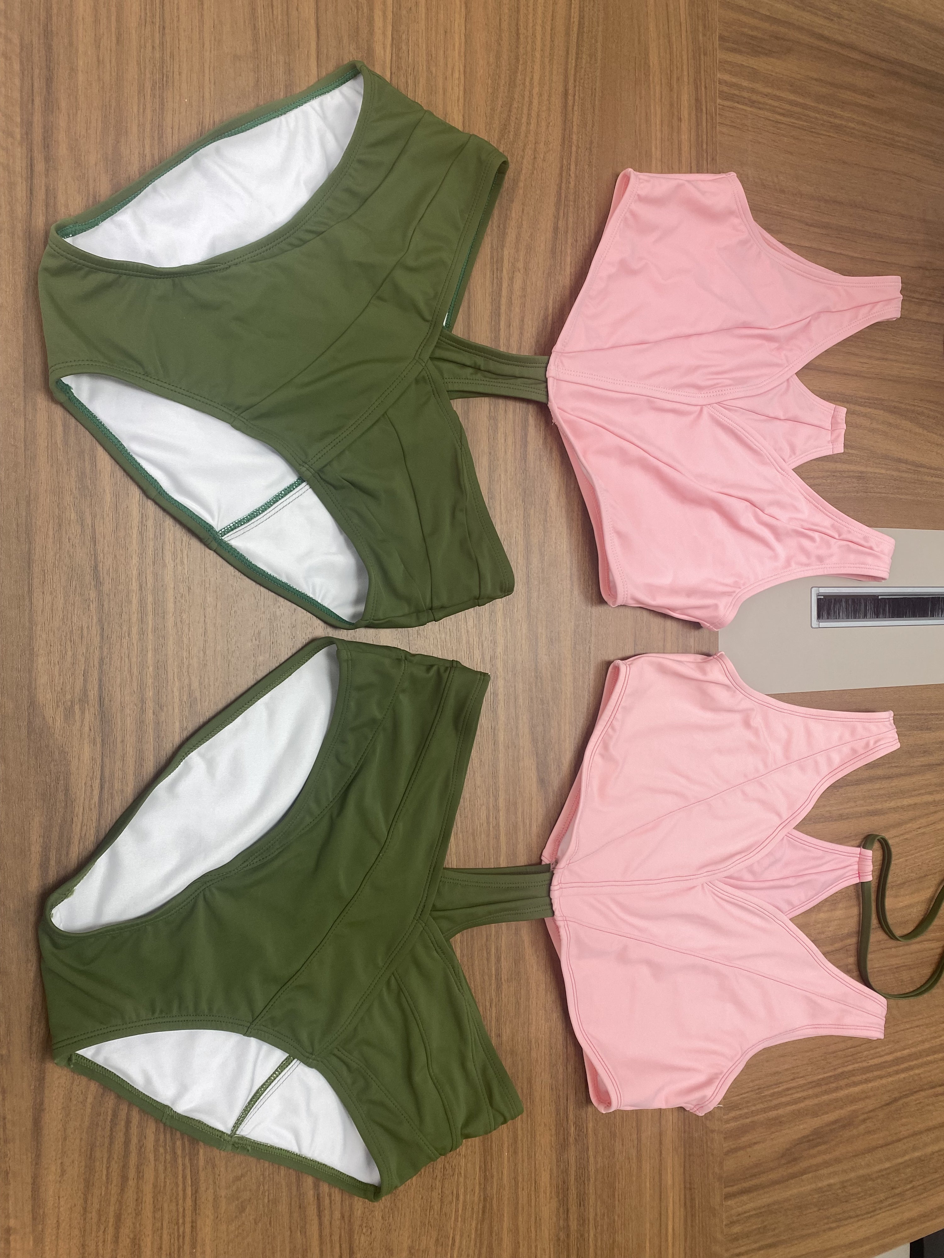 Two prototype sets of the Tulip Swimsuit laid flat on a wooden surface. Each set includes a green bottom shaped like overlapping leaves and a pink petal-shaped top. The suits are unassembled, showing separate front and back panels. The fabric has a soft matte finish, and the pieces are arranged side by side for comparison.