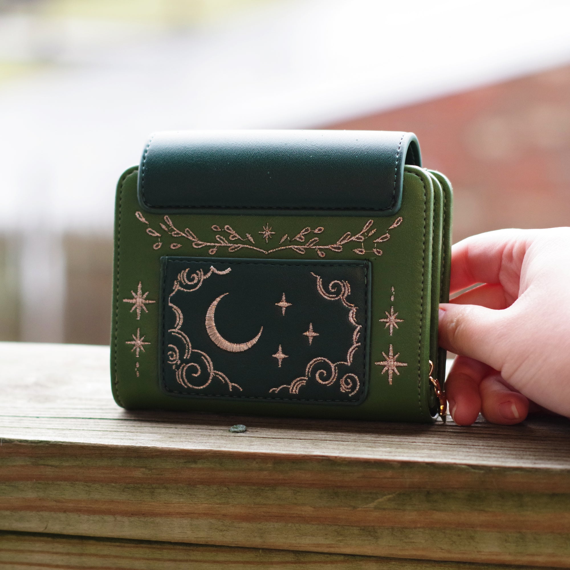 Green faux leather wallet with rose gold embroidery of a sleeping fox, floral vines, stars, and constellations. Held outdoors in natural lighting.