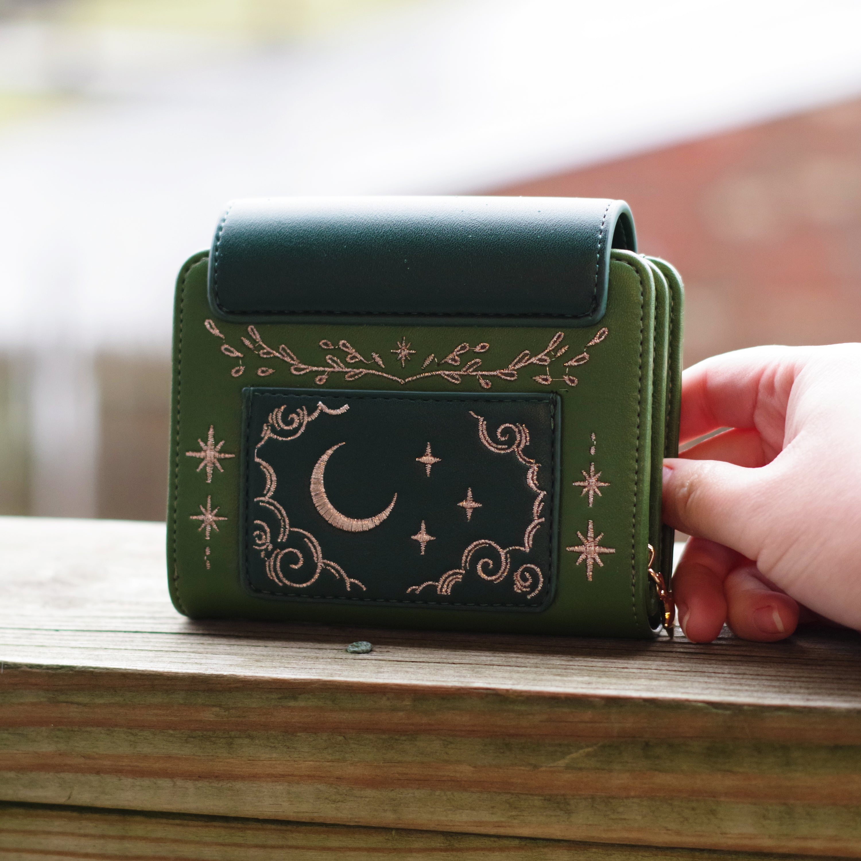 Green faux leather wallet with rose gold embroidery of a sleeping fox, floral vines, stars, and constellations. Held outdoors in natural lighting.