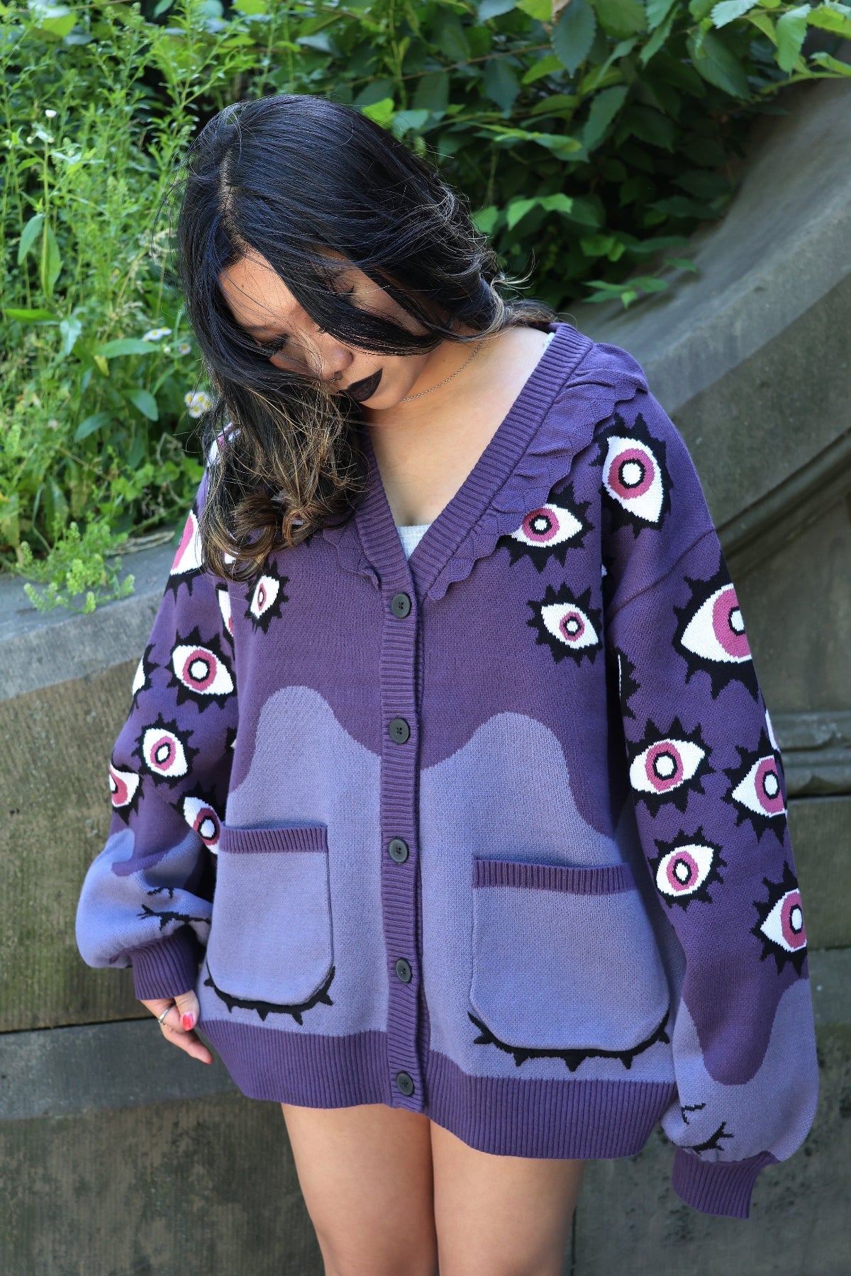 Model wearing the Abyss Cardigan, an oversized purple knit cardigan featuring eyeball motifs, gradient lavender details, and embroidered sleepy eye pockets, photographed outdoors in a garden setting.