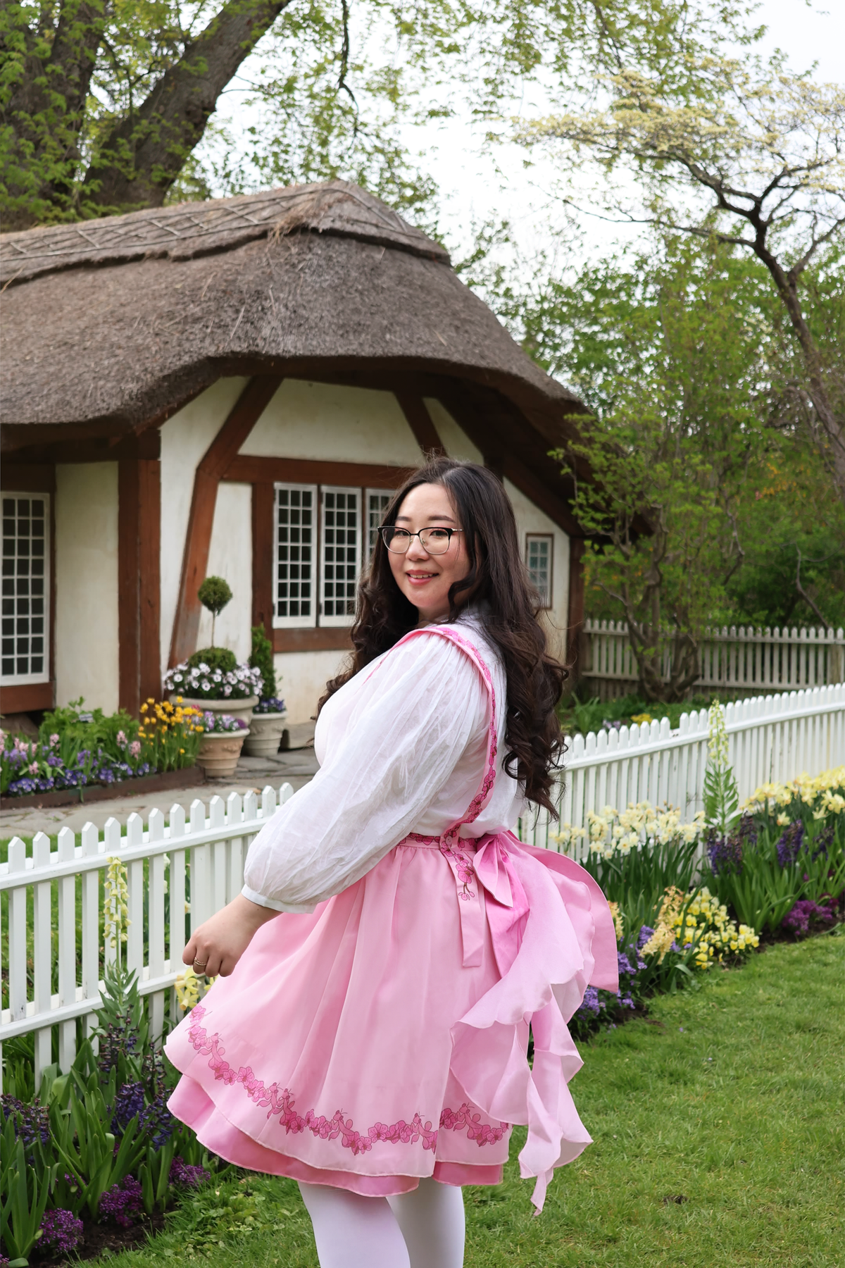 Model twirling in the Orchid Pinafore Skirt, a pink fairycore botanical-inspired pinafore dress with layered ruffles and floral lace trim in a cottage garden setting.