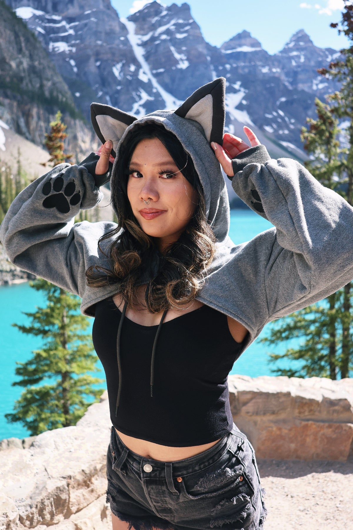 Model wearing the Nyahallo Wolf Shrug with wolf ears, embroidered paw print sleeves, and a cropped hoodie silhouette while adjusting the hood beside a turquoise mountain lake.