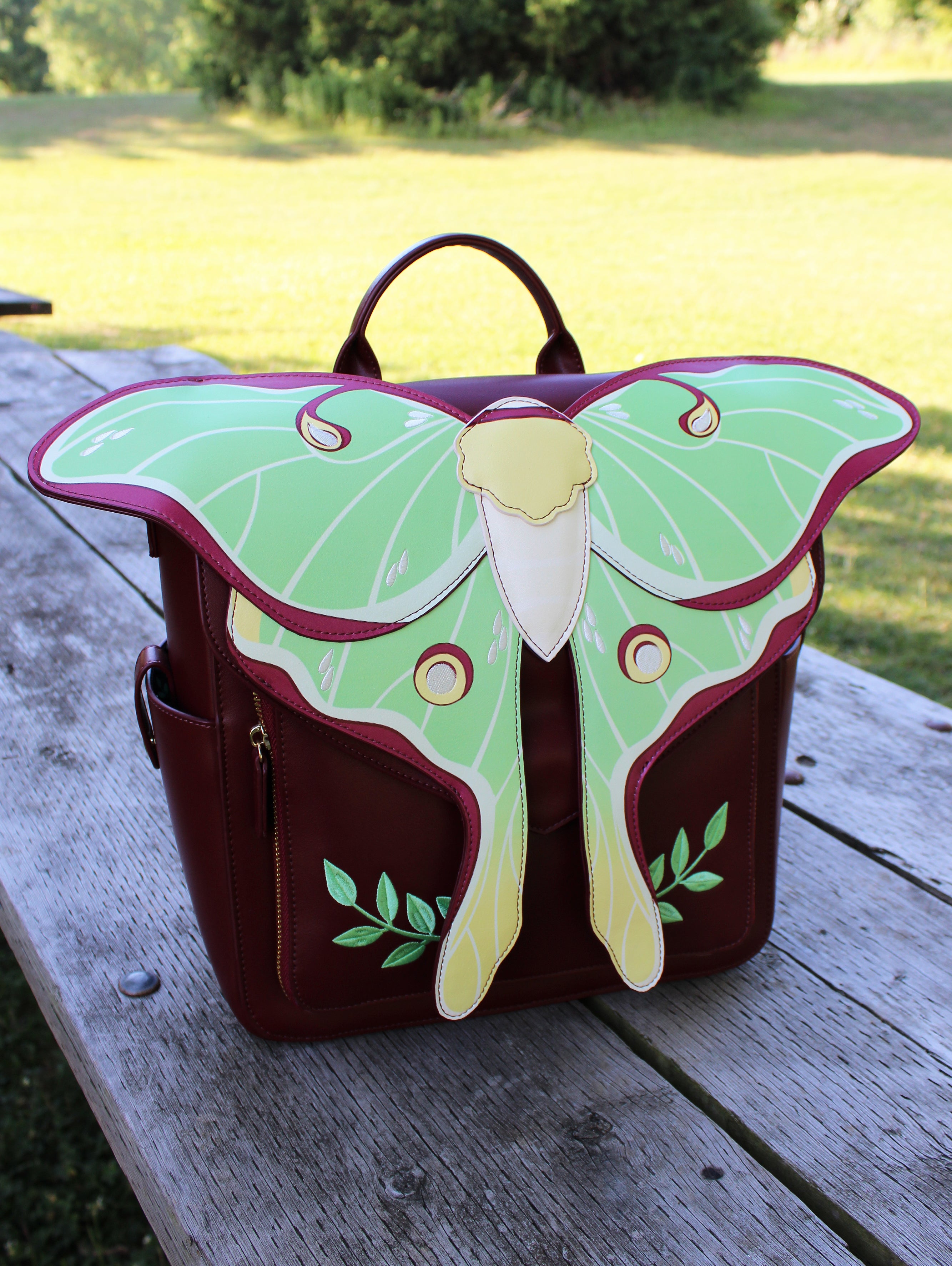 Burgundy backpack featuring a large green and yellow Luna moth design, photographed outdoors at a slight front angle on a weathered wooden picnic table.