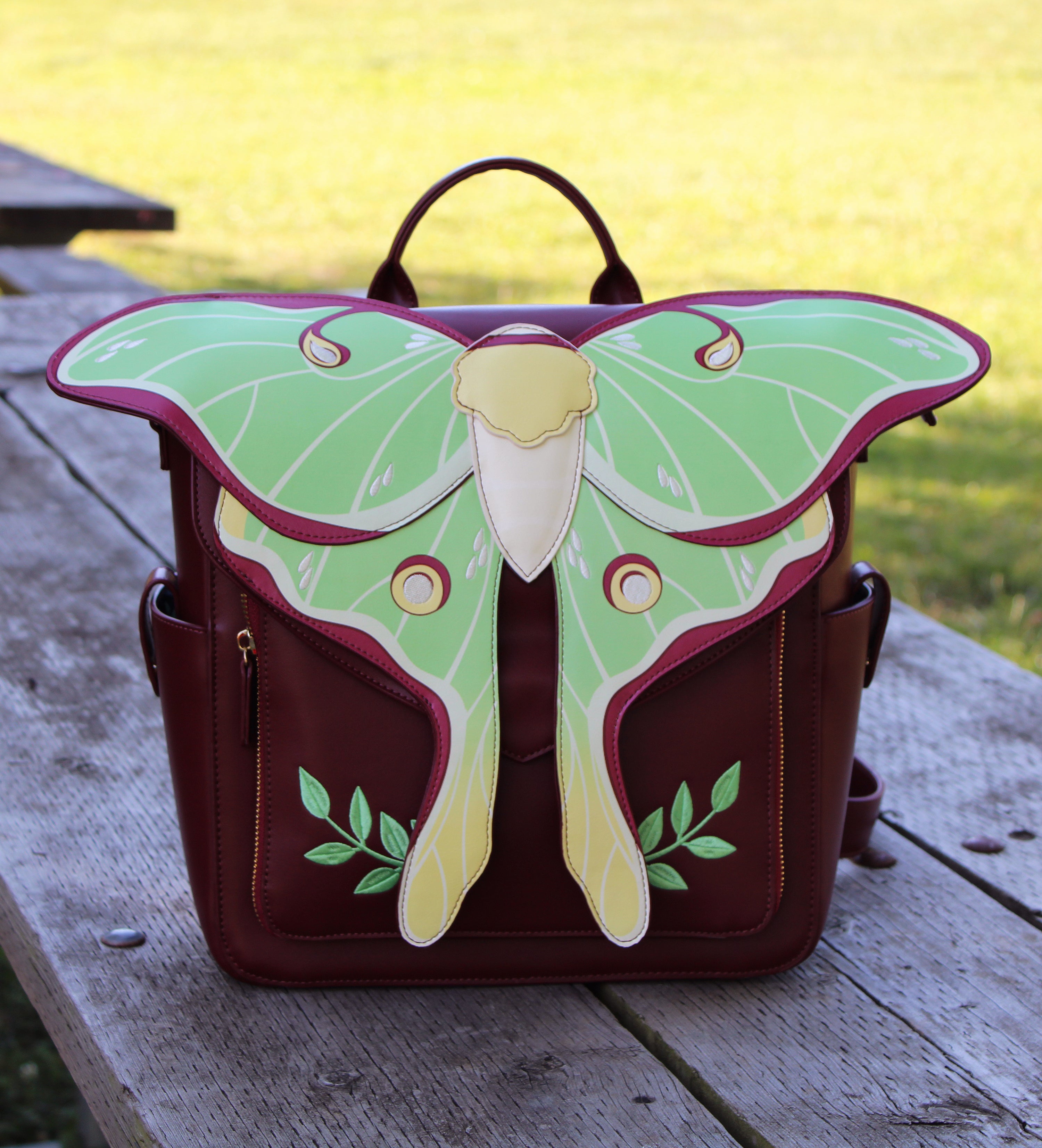 Burgundy backpack featuring a large green and yellow Luna moth design, photographed from the front while resting on a wooden picnic table outdoors.