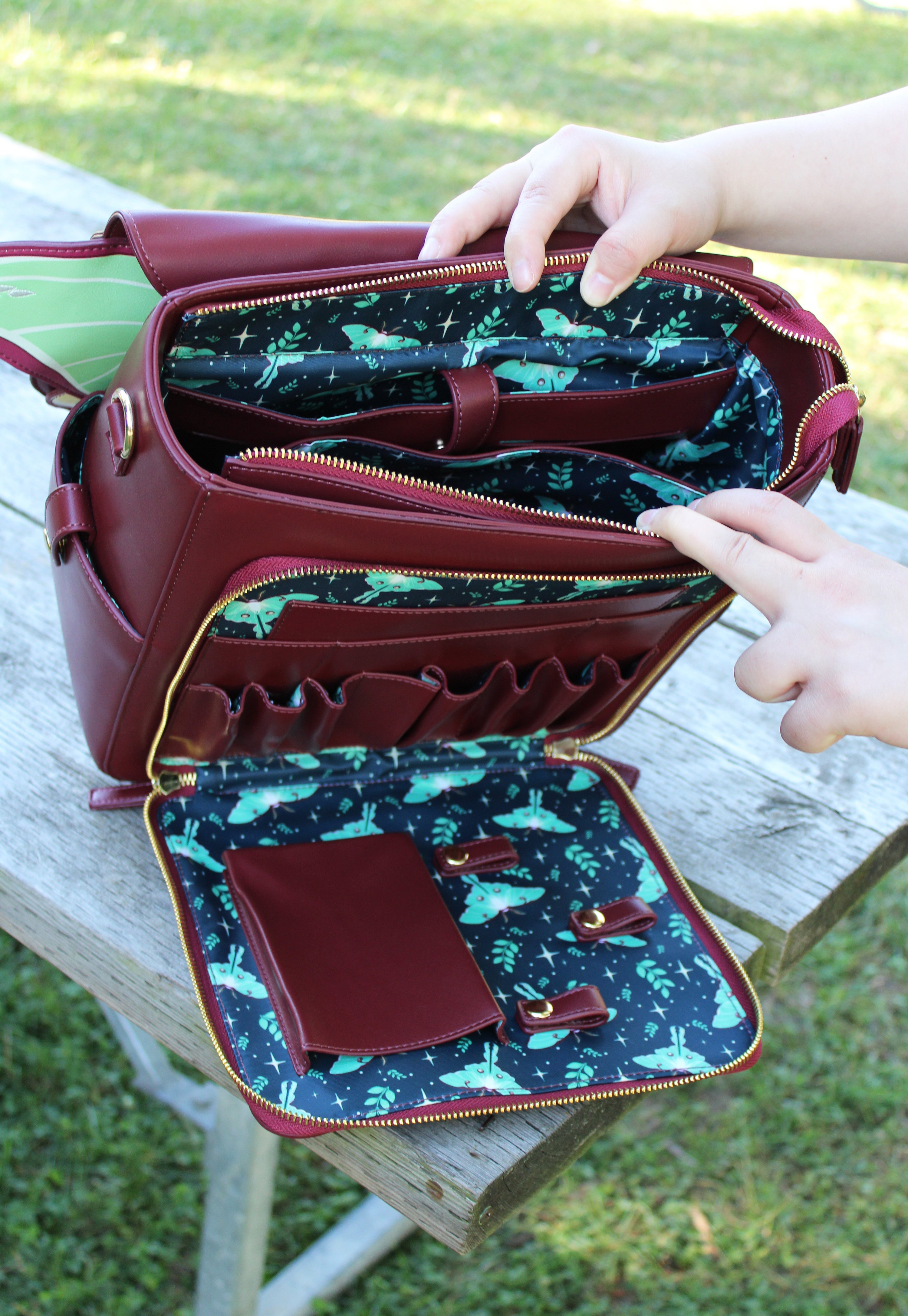 Open burgundy Luna moth backpack showing teal lining with moth and floral pattern, multiple interior pockets, and hands opening the bag on a wooden picnic table outdoors.