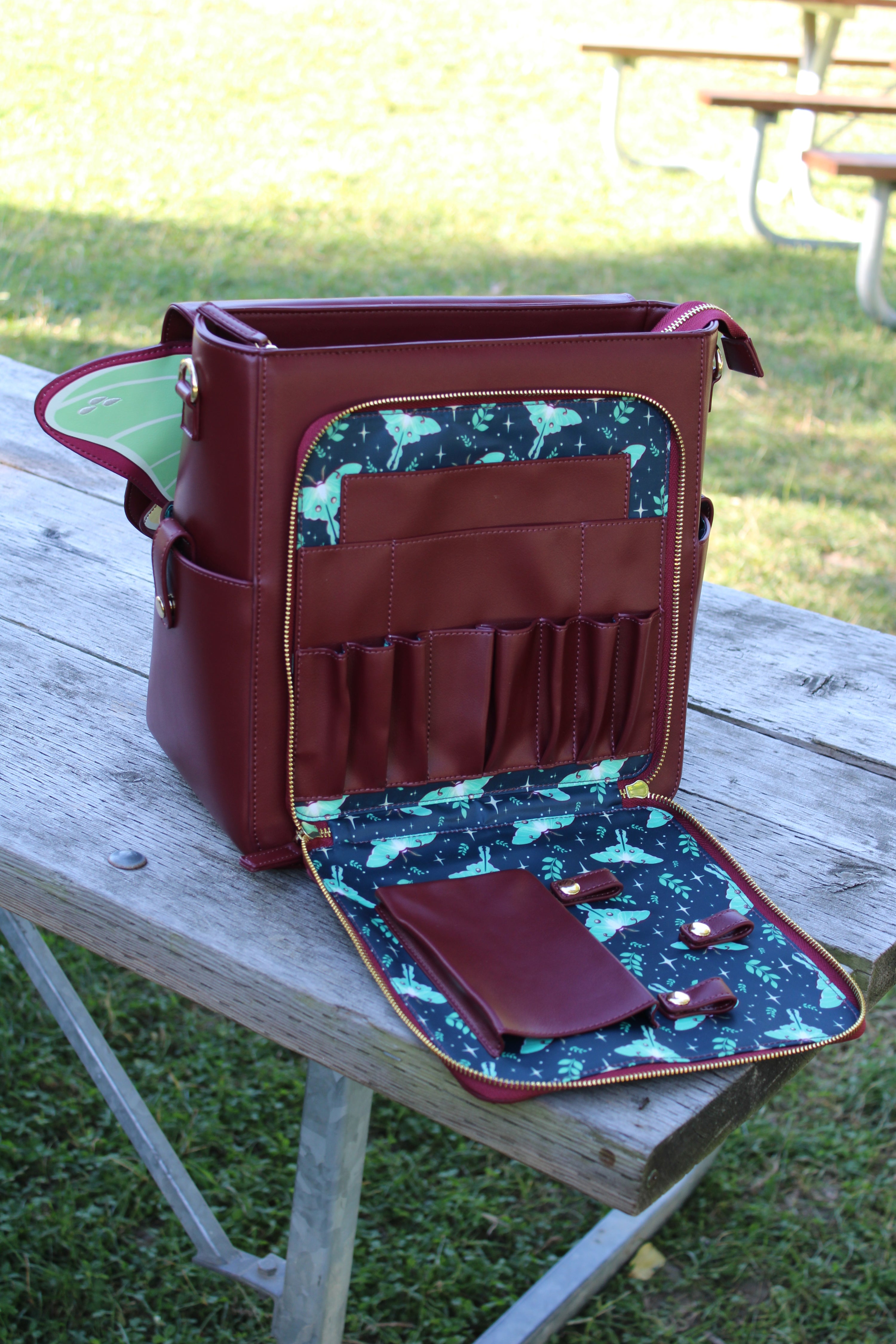 Open burgundy Luna moth backpack displayed outdoors on a wooden picnic table, showing teal lining with moth and floral pattern, multiple leather pockets, and an organizer panel with slots and loops.