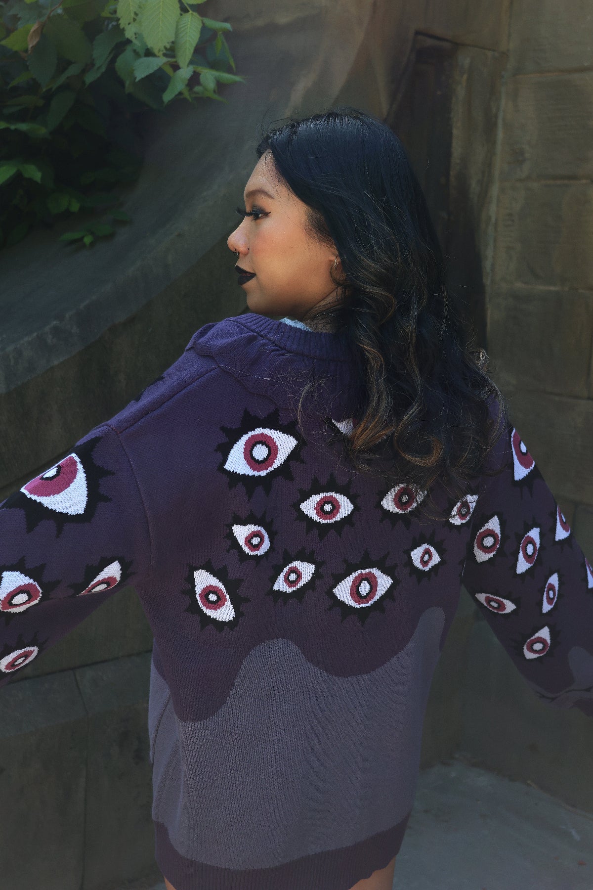 Back view of a model wearing the Abyss Cardigan, showcasing the oversized purple knit silhouette, gradient color blocking, and all-over pink and white eyeball pattern across the back and sleeves.