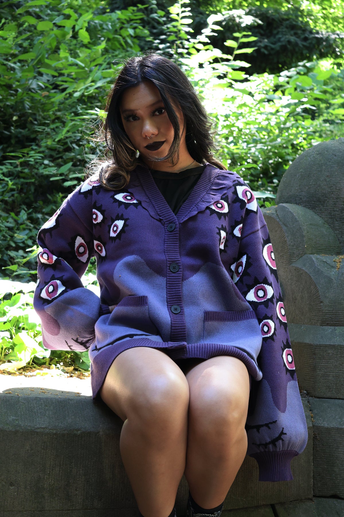 Model wearing the Abyss Cardigan, a purple oversized knit cardigan with all-over eyeball motifs, seated outdoors in a lush forest setting.
