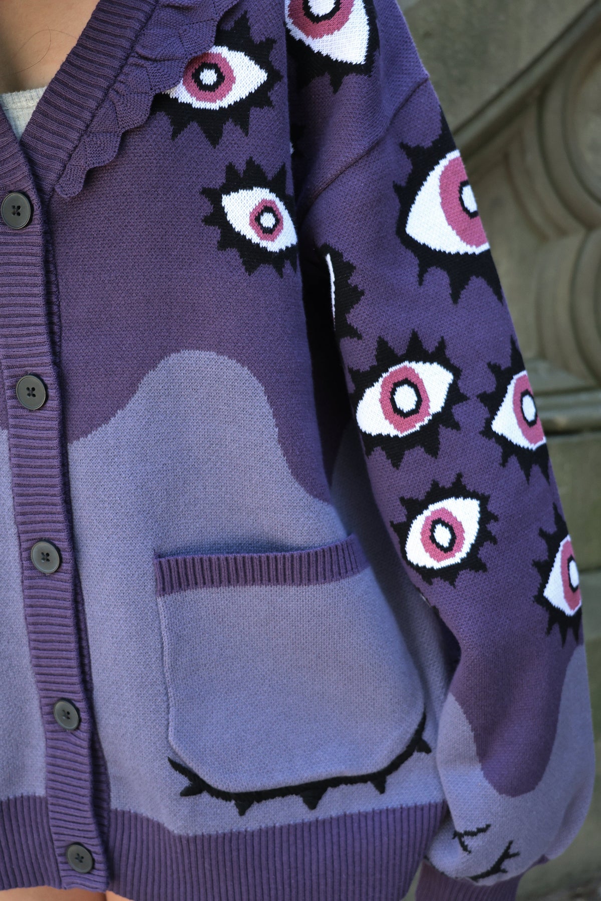 Close-up detail of the Abyss Cardigan featuring purple knit fabric, all-over eyeball motifs on the sleeves, button-front closure, and embroidered sleepy eye pocket accents.