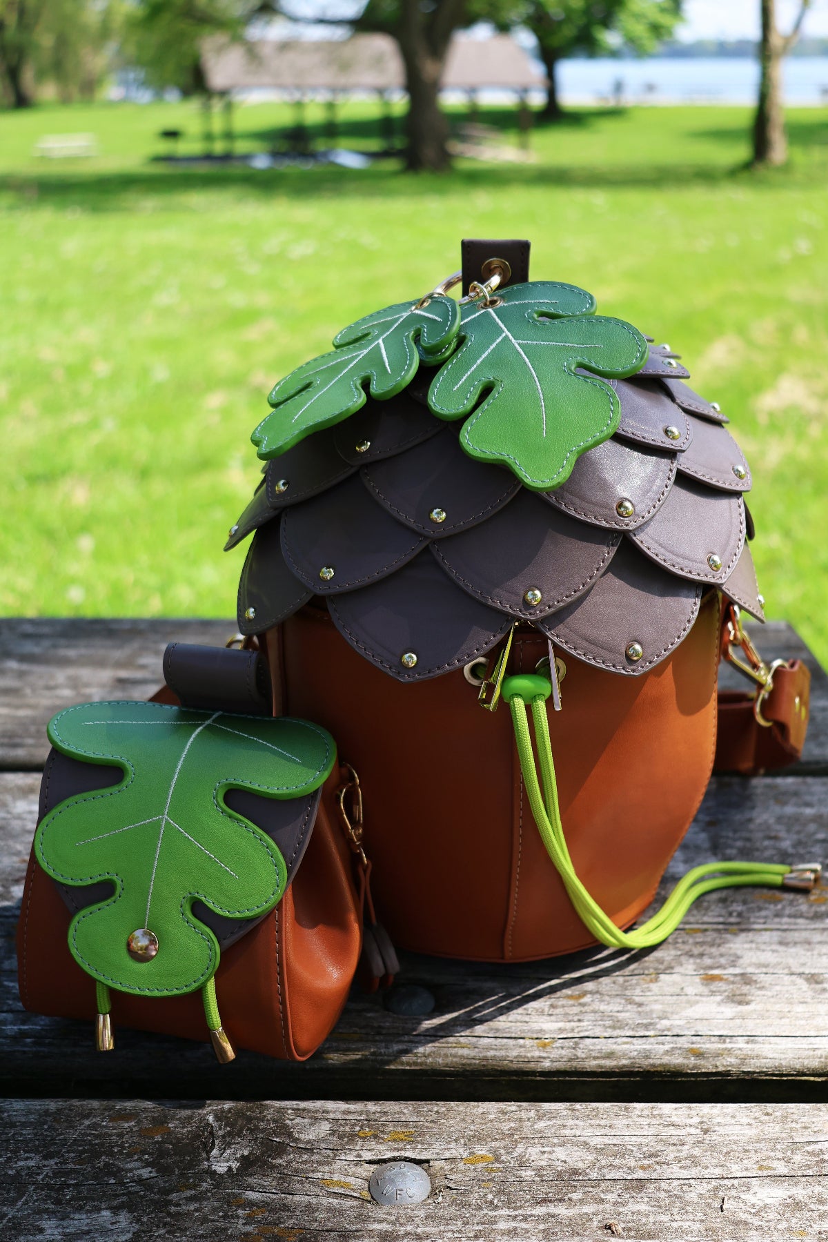 Acorn-shaped brown crossbody bag with layered cap details and oak leaf charms displayed beside a matching oak leaf coin pouch on a wooden picnic table.