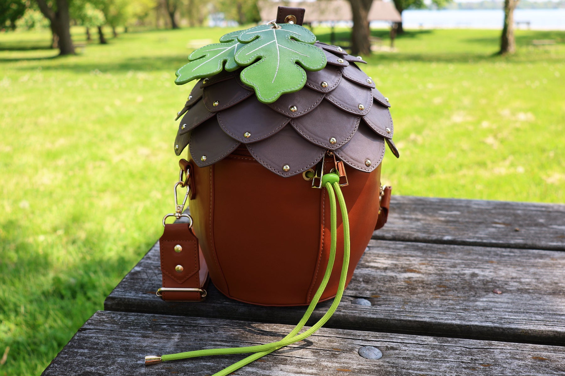 Acorn-shaped brown crossbody bag with layered acorn cap details, oak leaf accents, gold hardware, and green drawstring closure displayed outdoors on a wooden picnic table.