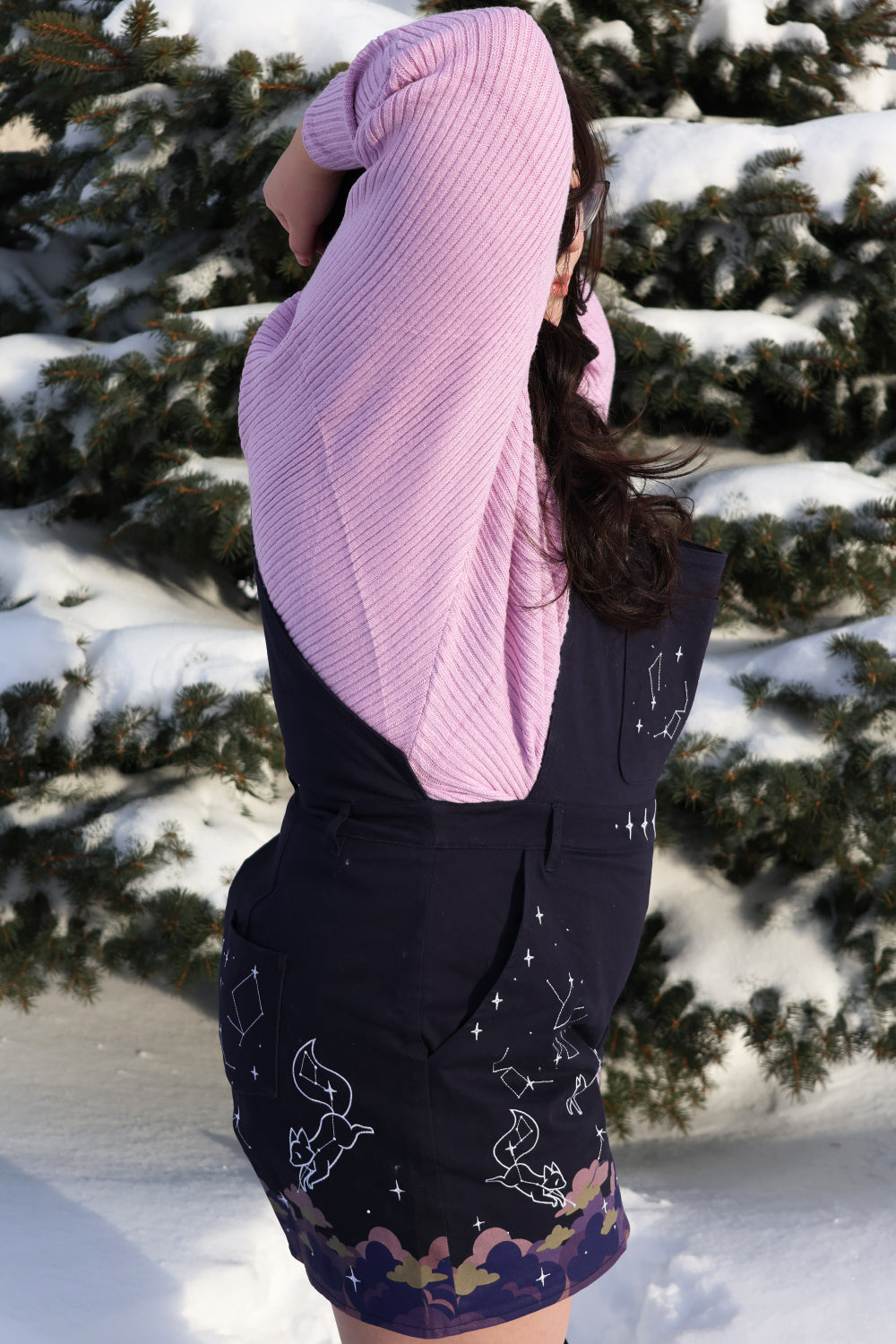 Person modeling navy celestial themed overalls with constellation prints and pastel cloud details beside snow covered pine trees.