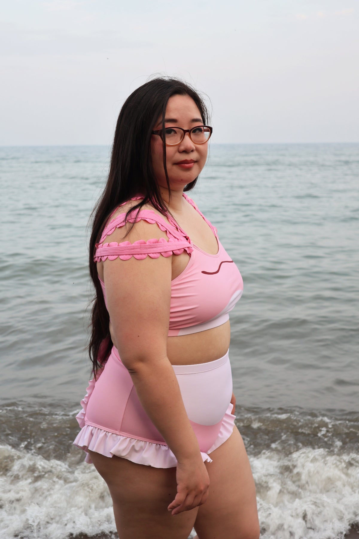 Side view of a model wearing the pink Axolotl two-piece swimsuit, featuring decorative axolotl-inspired shoulder details and high-waisted ruffle-trim bottoms at the beach.
