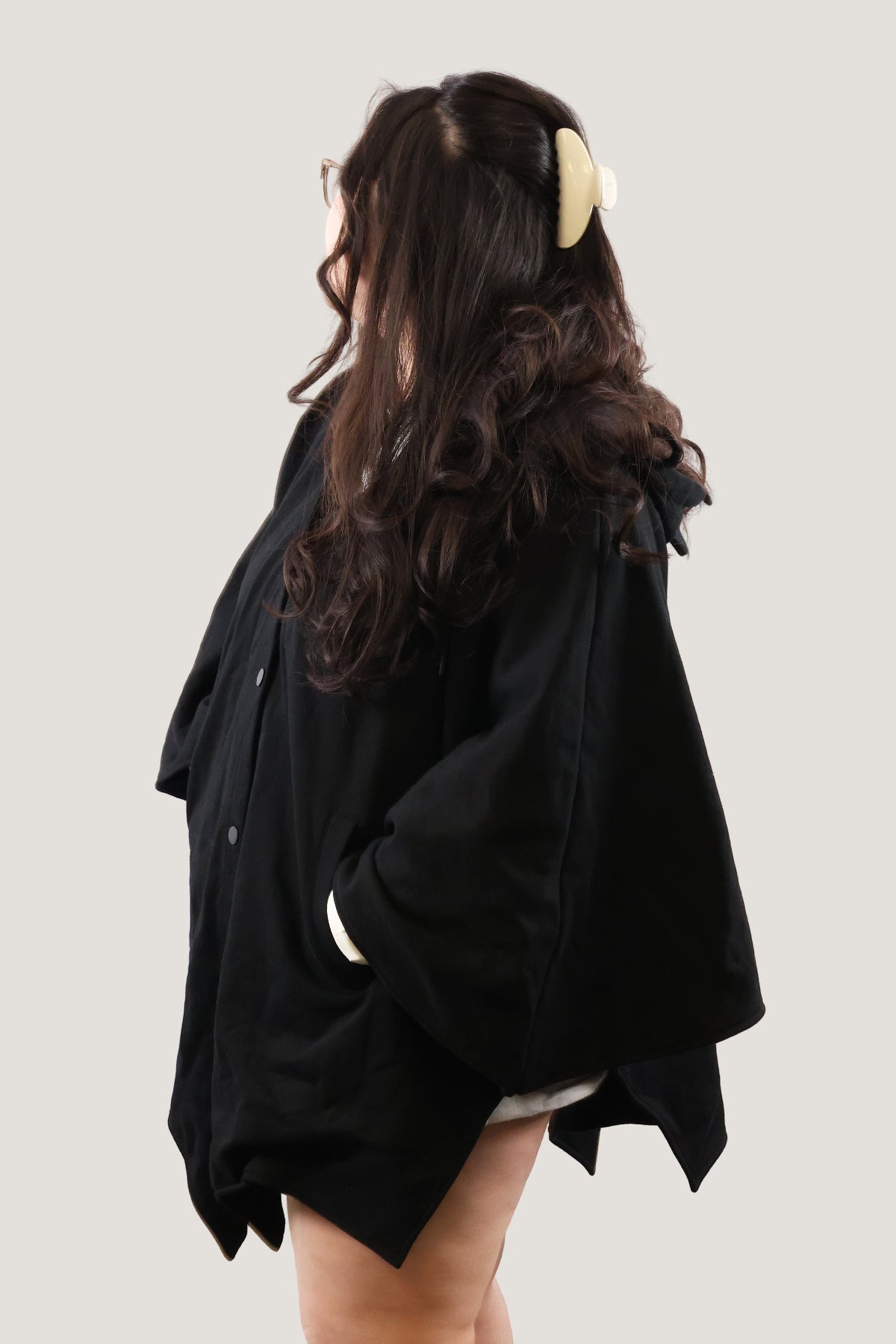Back angled view of model wearing a black bat-inspired hooded cloak with hands in pockets, showing draped silhouette against a light background