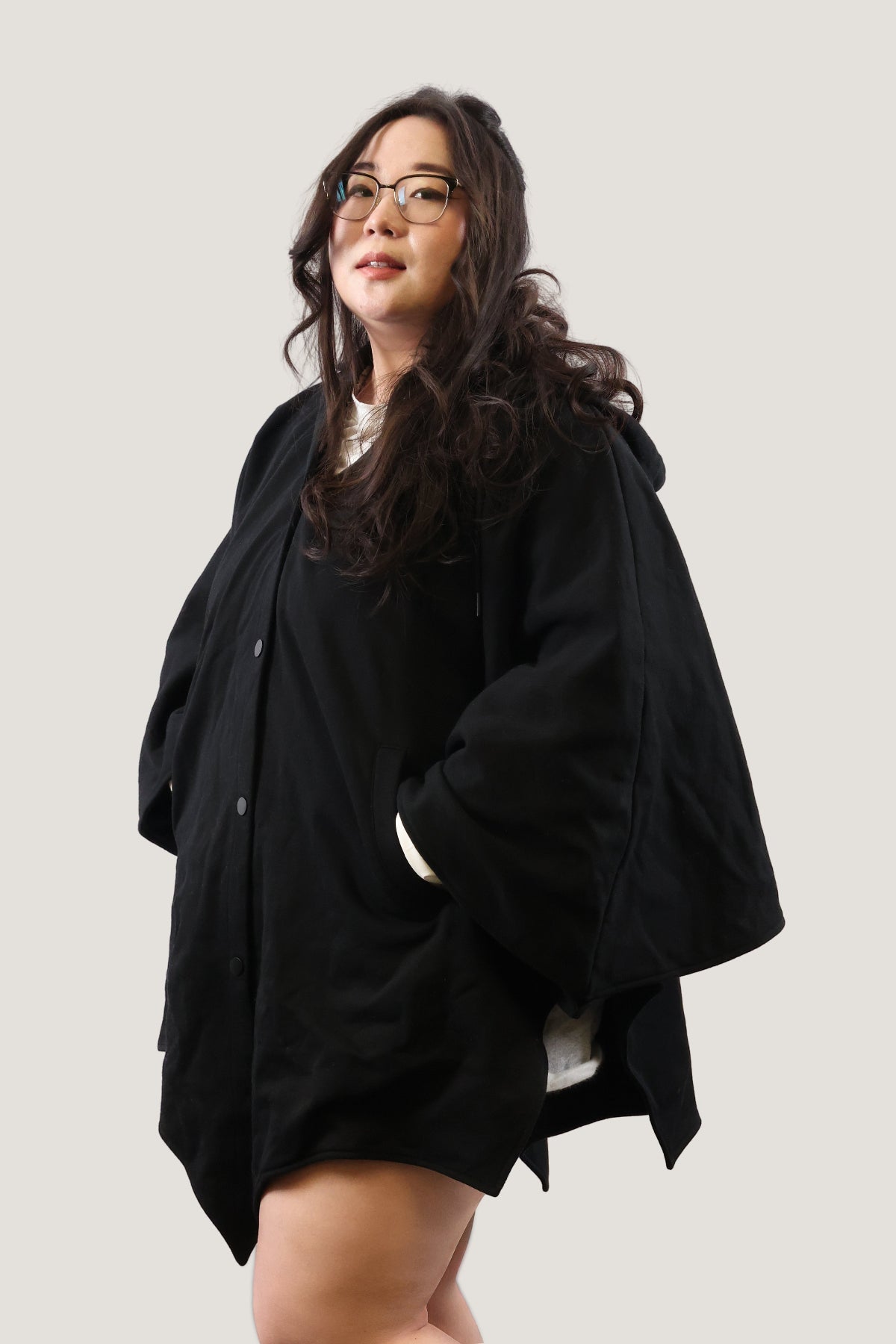 Model wearing a black bat-inspired hooded cloak with hands in pockets, showing relaxed oversized fit and everyday styling against a light background