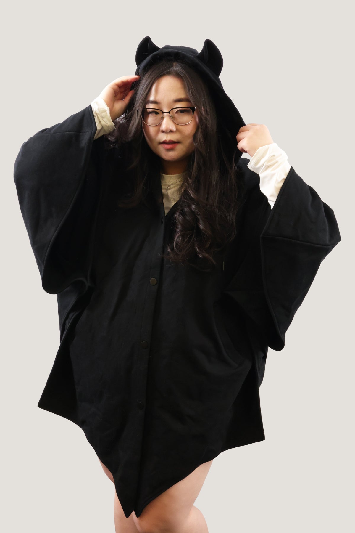 Front view of model adjusting the hood of a black bat-inspired cloak with pointed ear details, showing oversized fit and draped sleeves against a light background