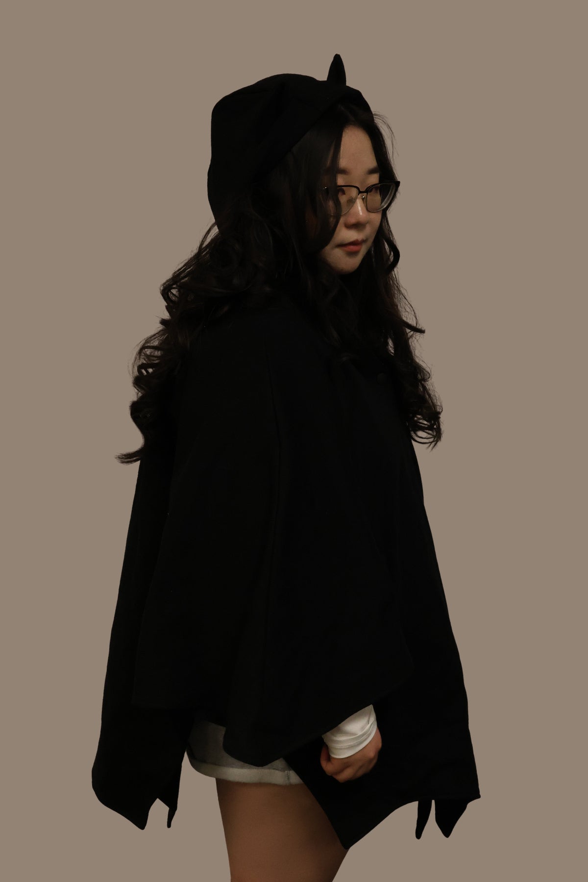 Side view of model wearing a black bat-inspired hooded cloak with pointed ear details, shown in a darker toned setting against a neutral background