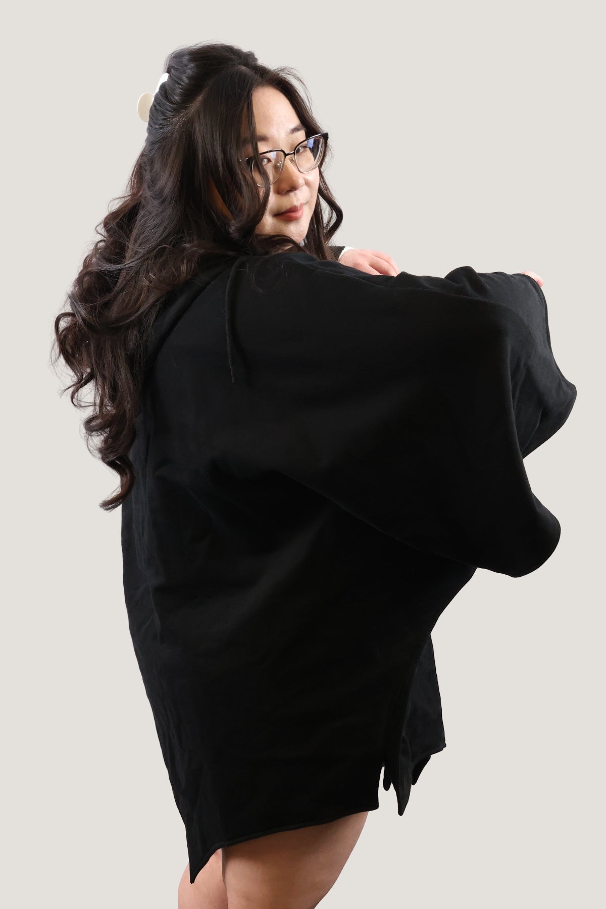 Over-the-shoulder back view of model wearing a black bat-inspired hooded cloak, highlighting the smooth drape and relaxed silhouette against a light background