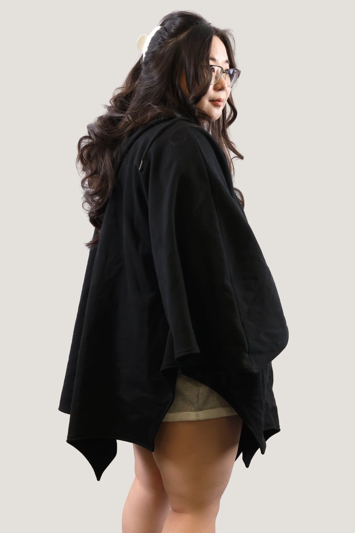 Side back view of model wearing a black bat-inspired hooded cloak, highlighting the draped silhouette and pointed hem against a light background