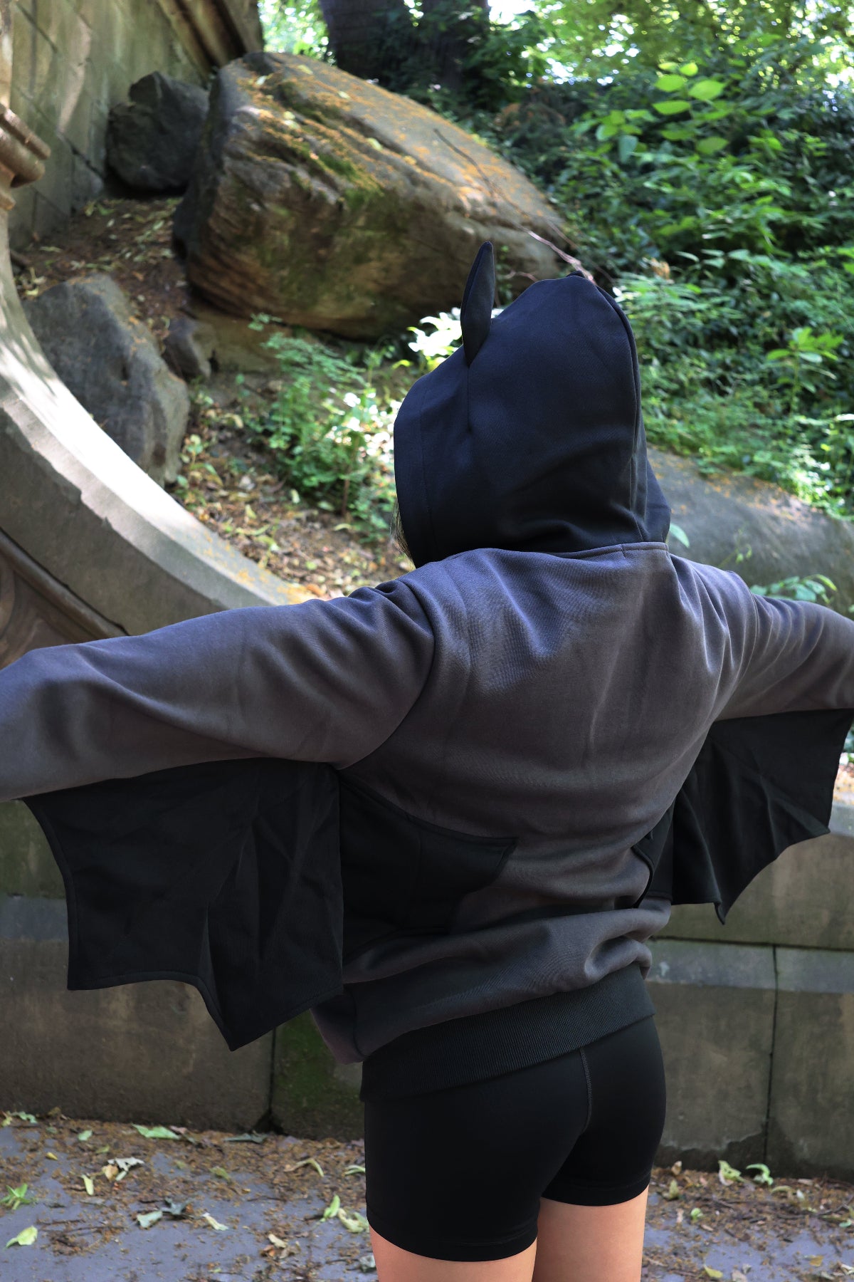 Back view of a model wearing a black bat-inspired hoodie with pointed bat ears and extended bat wing sleeves in an outdoor forest setting.