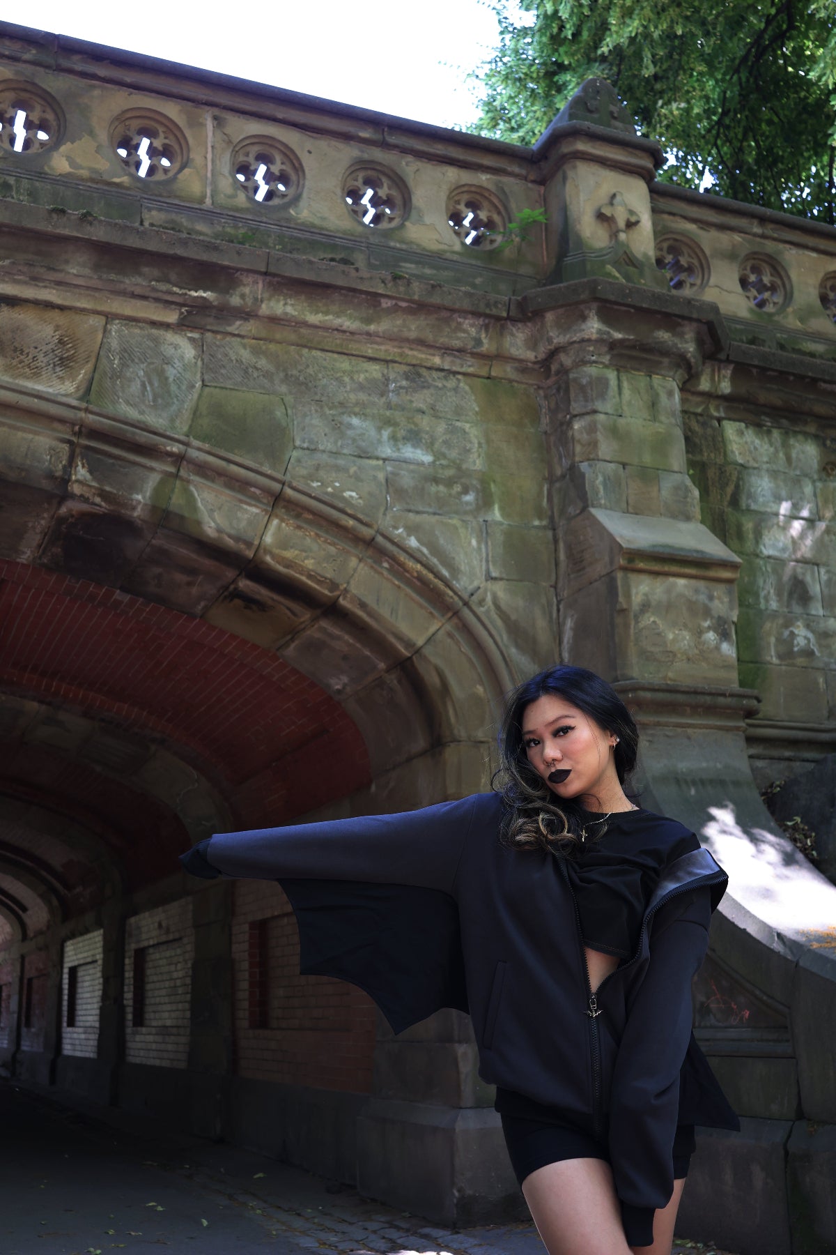Model wearing a black bat hoodie with extended bat wing sleeves, posing beside a stone archway in a park to showcase the wing-inspired silhouette and gothic design details.