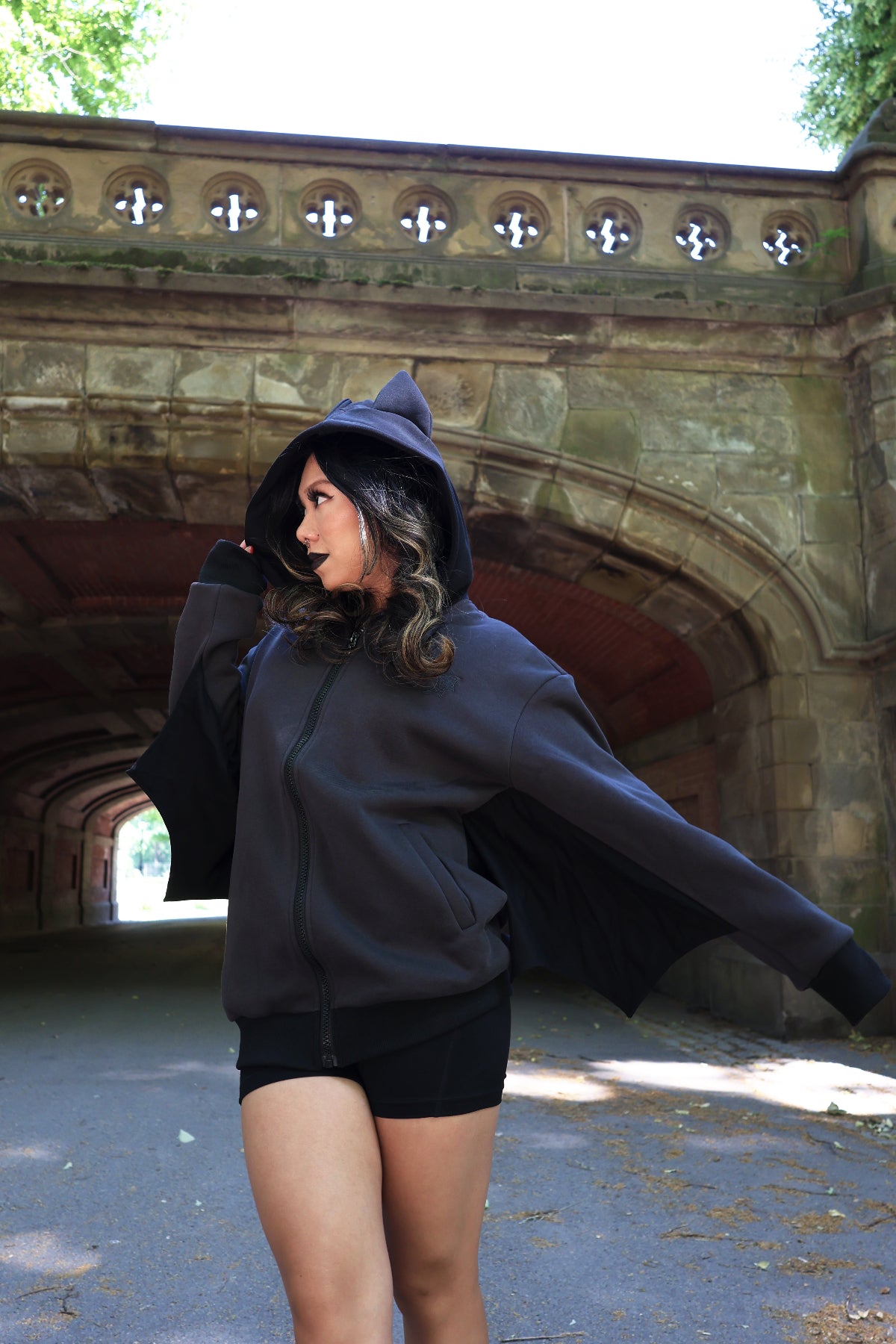 Model wearing a black bat-inspired hoodie with pointed bat ears and bat wing sleeves, posing beneath a stone archway outdoors.