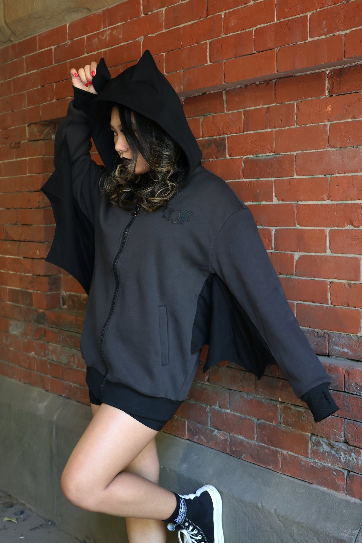 Model wearing a black bat-inspired hoodie with pointed bat ears and bat wing sleeves, posing against a red brick wall.