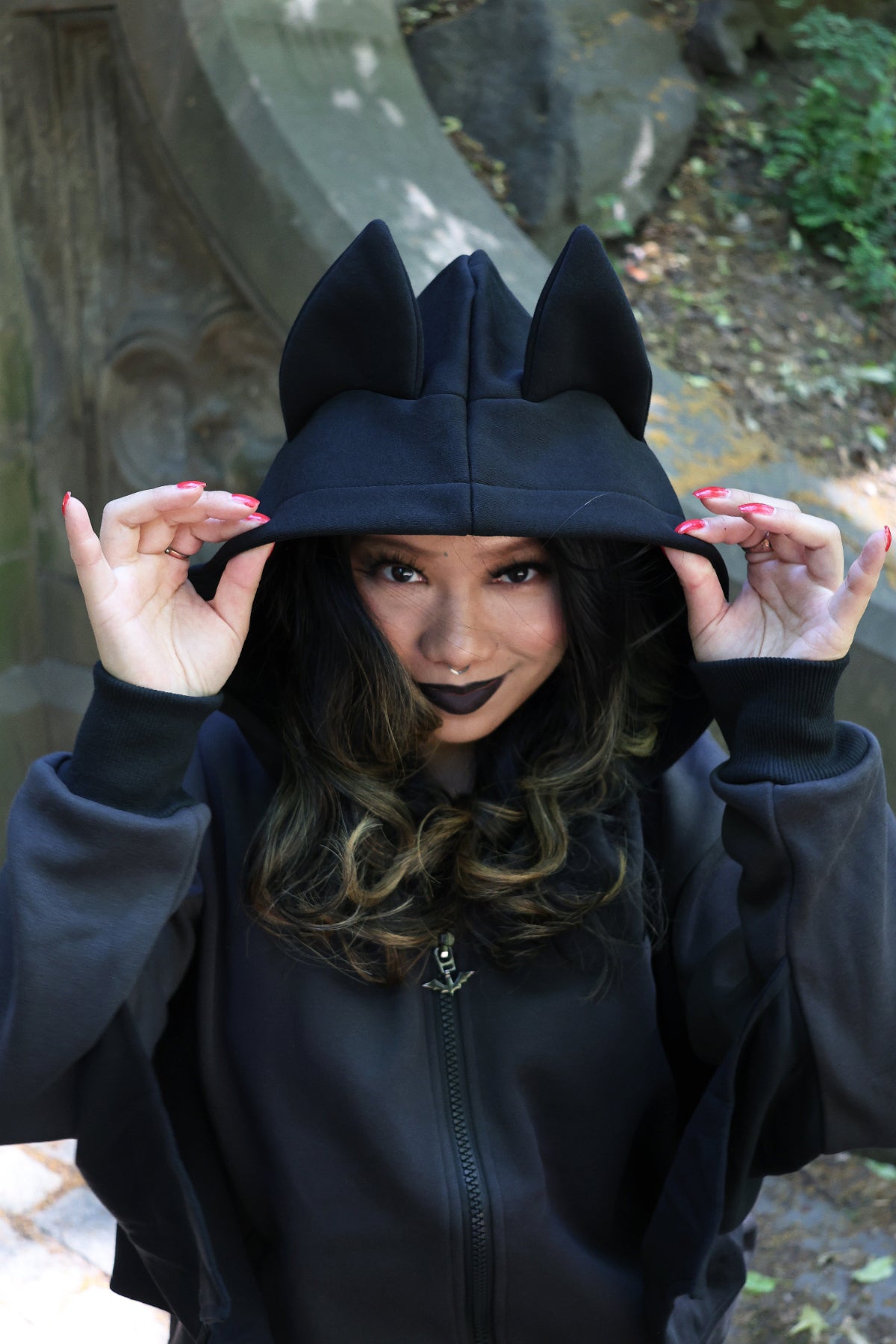 Model wearing a black bat-inspired hoodie with pointed bat ears on the hood, holding the hood edges in an outdoor setting.