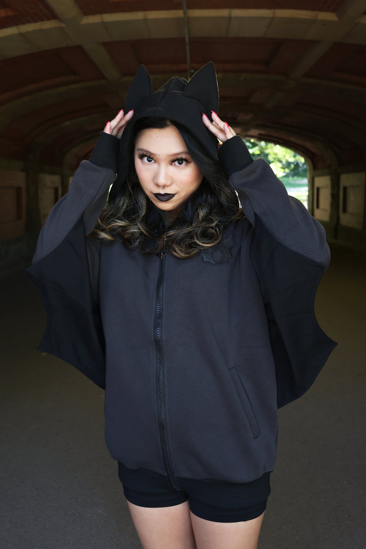 Front view of a model wearing a black bat-inspired hoodie with pointed bat ears and dramatic bat wing sleeves, standing beneath a stone tunnel outdoors.