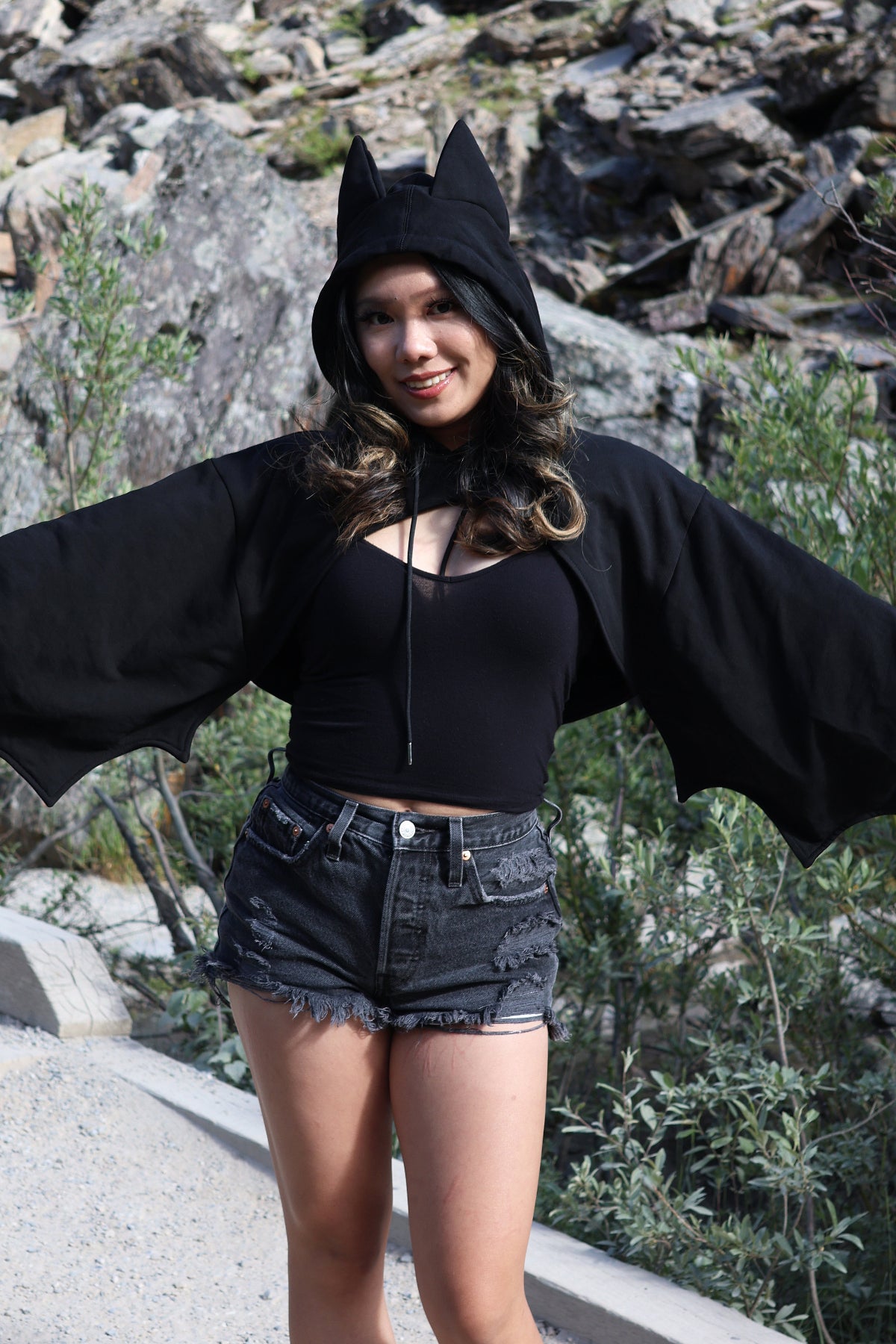 Model wearing the Nyahallo Bat Shrug with bat ears on the hood and oversized bat wing sleeves, posing with arms outstretched to showcase the wing-inspired silhouette.