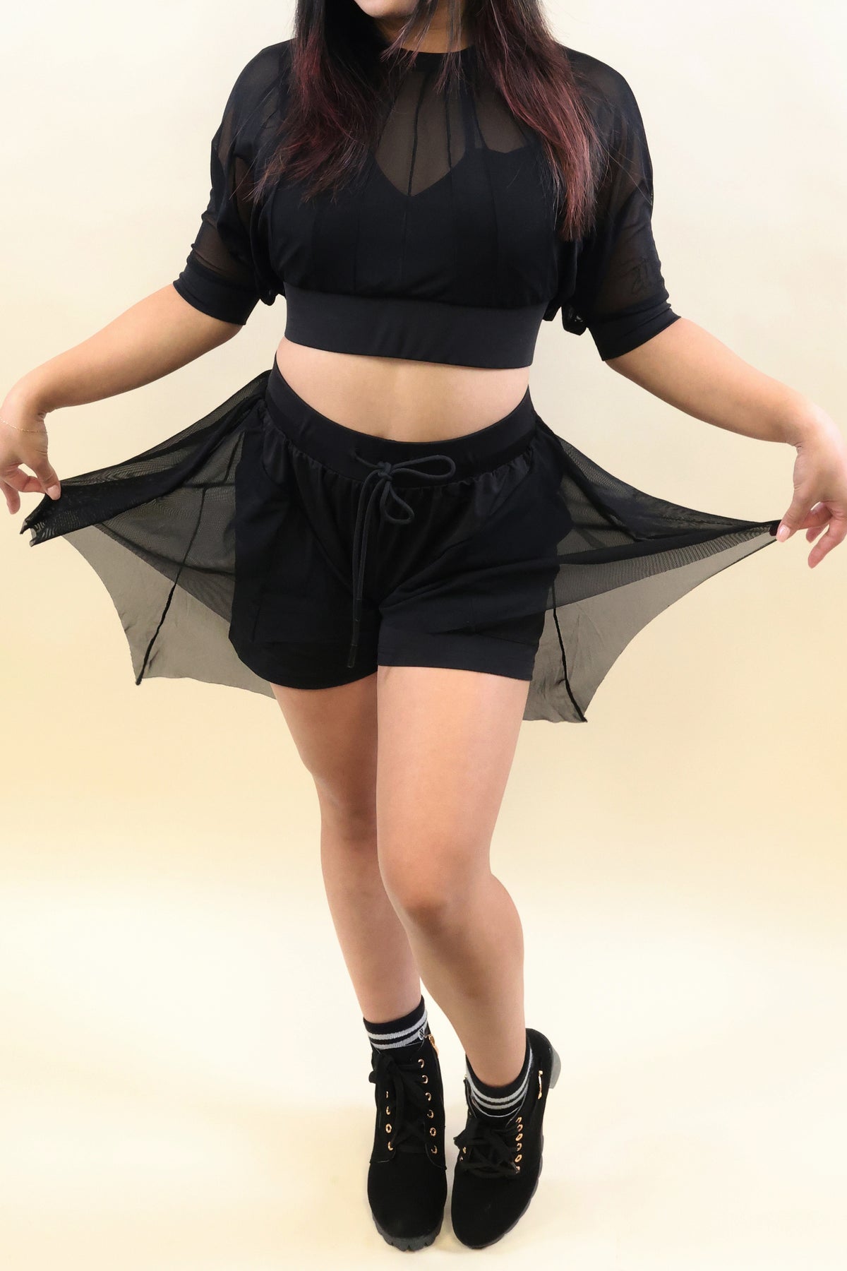 A model wearing a black sheer mesh two-piece outfit consisting of a short-sleeve crop top and matching drawstring shorts, holding the sides of the skirted shorts to show the sheer fabric. The model is also wearing black lace-up boots and striped socks, set against a plain beige studio background.