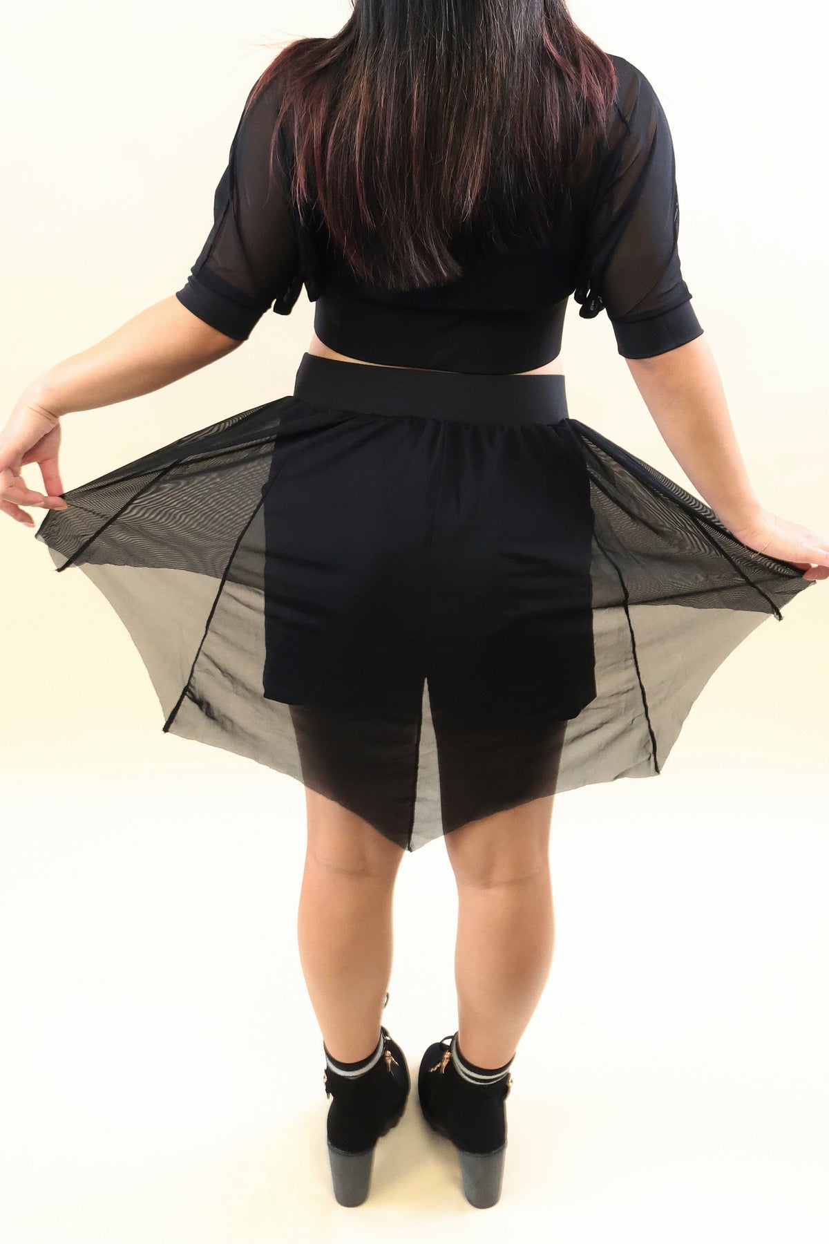 A model shown from the back wearing a black sheer mesh crop top and matching sheer mesh shorts, holding out the sides of the skirt to show its volume, against a plain light background.