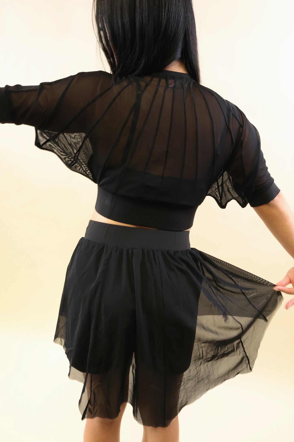 A model shown from the back wearing a black sheer mesh two-piece outfit with a cropped long-sleeve top and matching high-waisted shorts, holding out the skirt to show its flow against a plain light beige background.