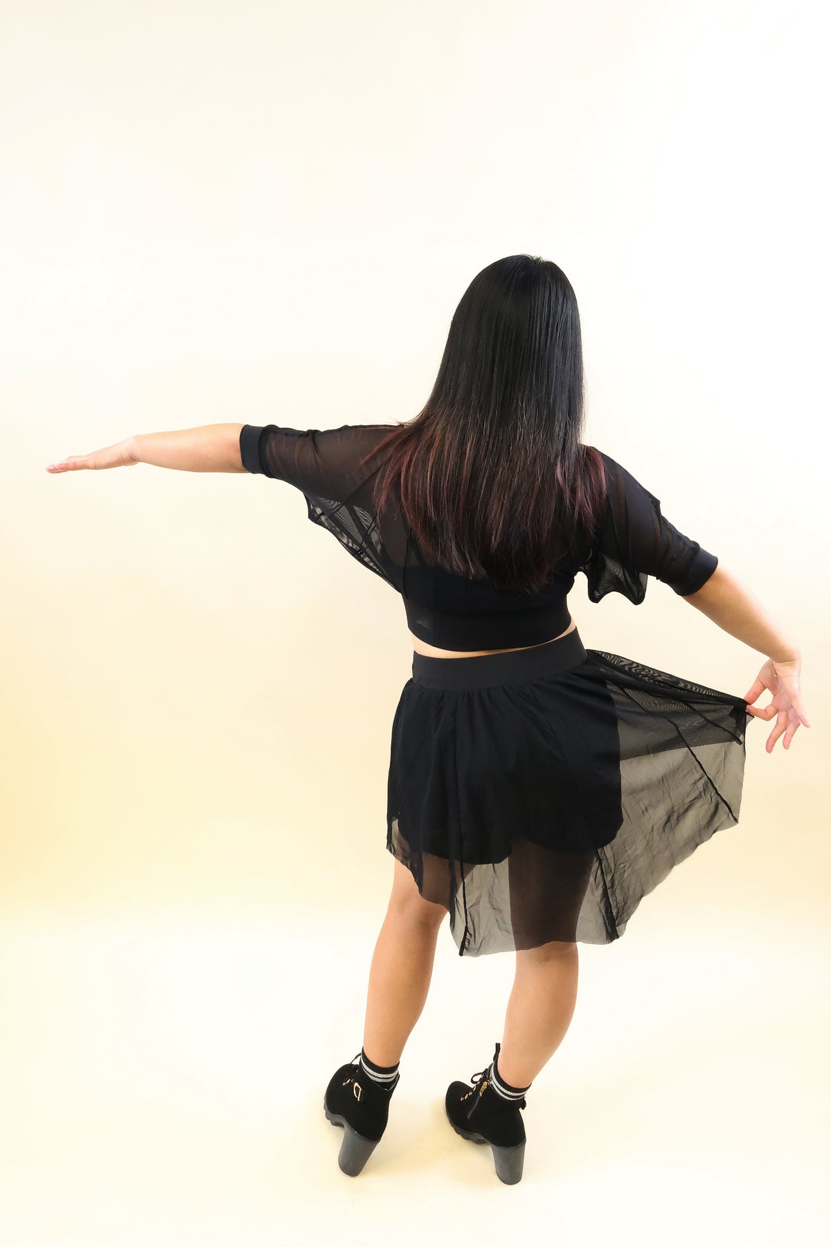 A woman with long dark hair is shown from behind wearing a black sheer long-sleeve crop top and a matching black sheer mini skirt, holding out her arms and lifting the skirt slightly to show movement. She is wearing black heels against a plain light beige studio background.