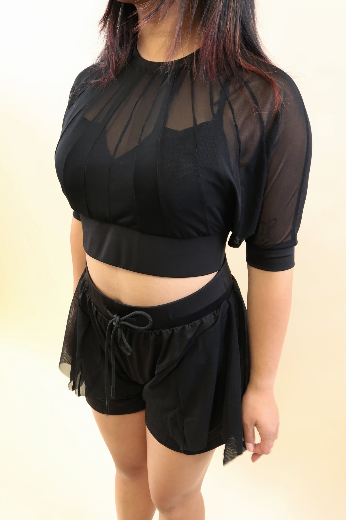 A woman wearing a black sheer mesh crop top with elbow-length sleeves and matching black drawstring shorts, photographed against a plain light beige studio background.