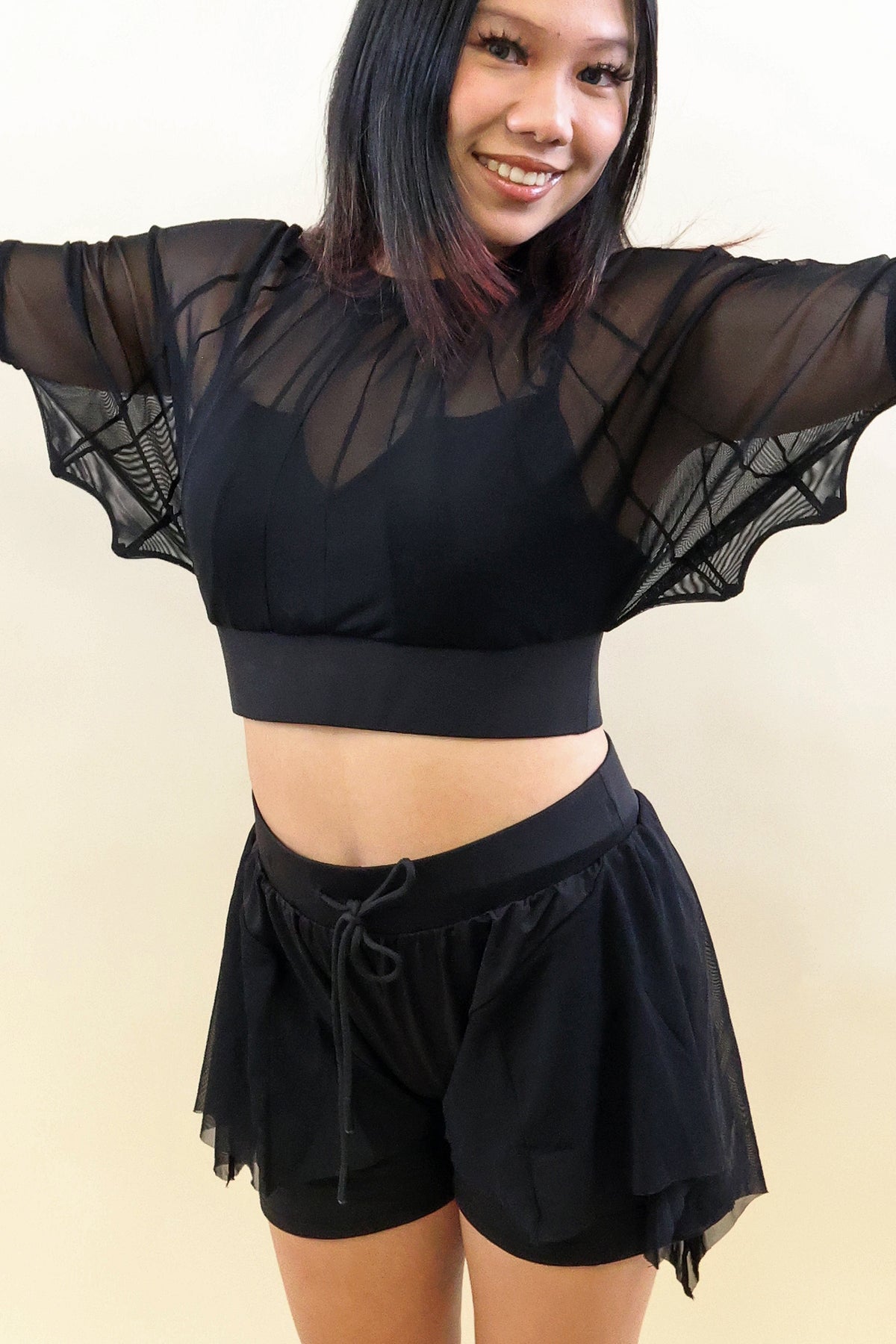A woman modeling a black sheer mesh crop top with dramatic wide sleeves and matching black drawstring shorts against a plain beige background.