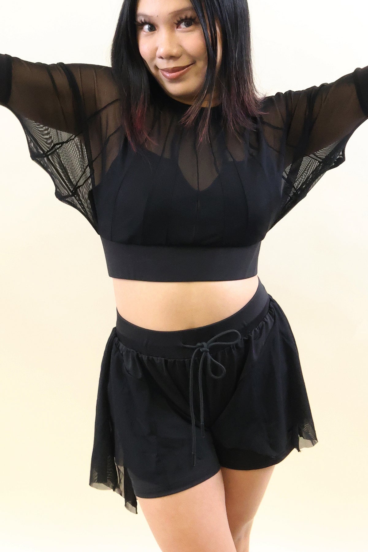 A woman modeling a black two-piece activewear set featuring a sheer long-sleeve crop top and matching drawstring shorts against a plain light beige background.