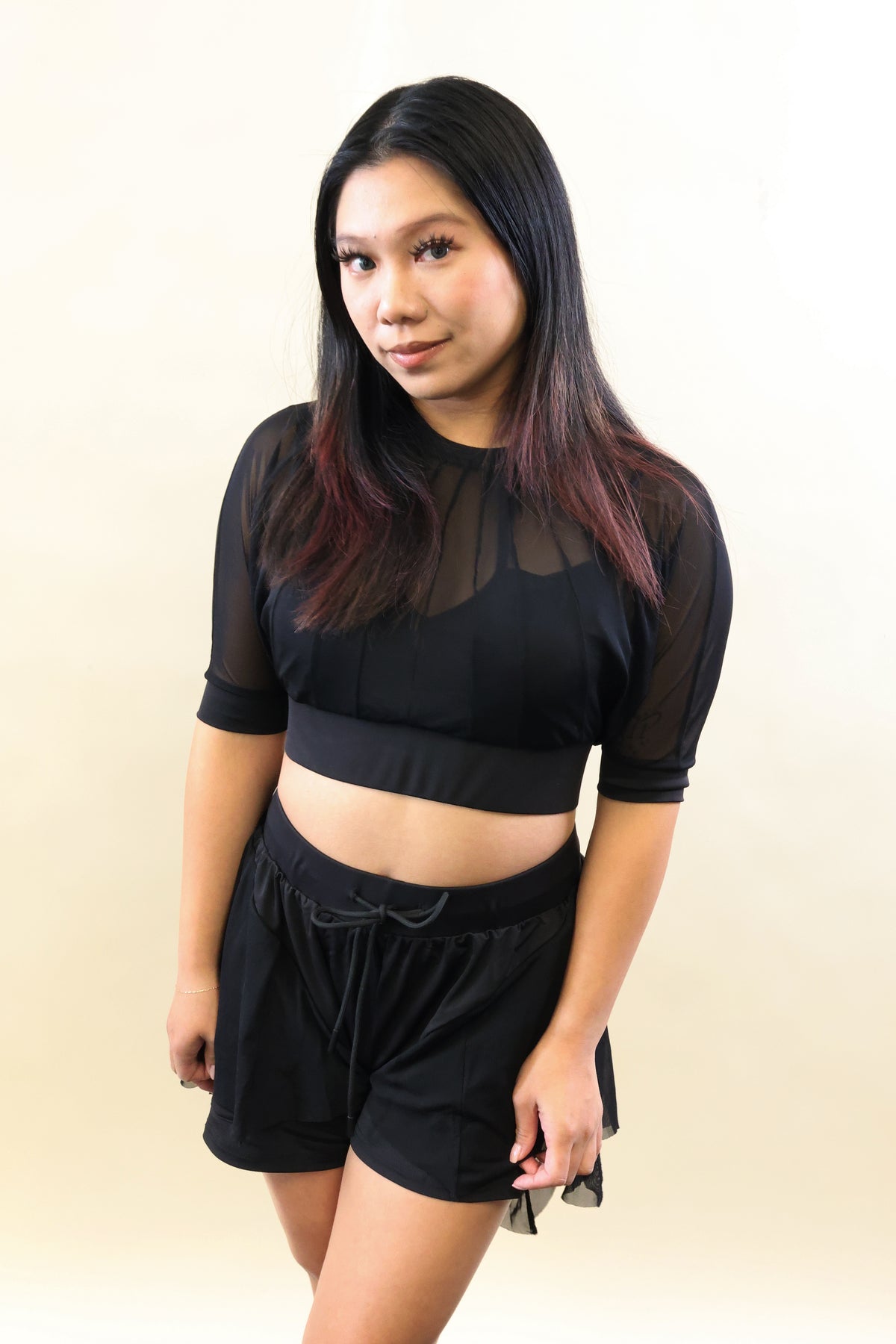 A woman modeling a black sheer mesh short-sleeve crop top and matching black shorts against a plain beige studio background.