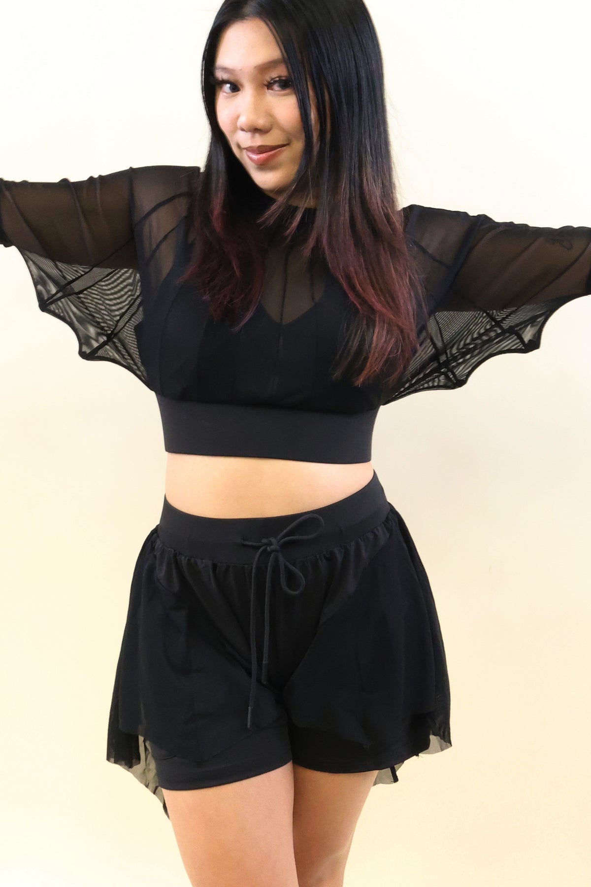 A woman modeling a black two-piece outfit consisting of a sheer mesh crop top with dramatic winged sleeves and matching black drawstring shorts, set against a plain light beige background.