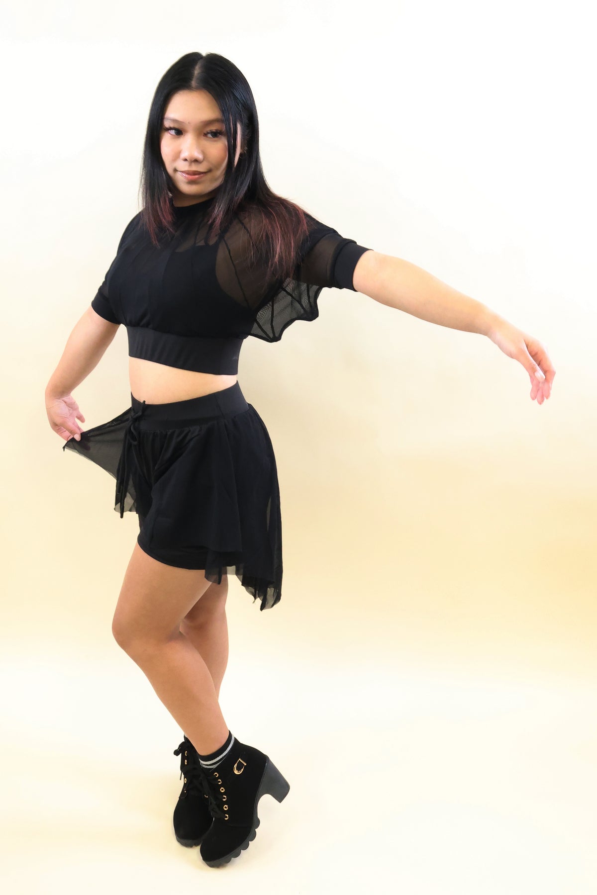 A woman modeling a black two-piece outfit consisting of a sheer short-sleeve crop top and a matching black mini skirt, paired with black lace-up ankle boots, against a plain cream background.