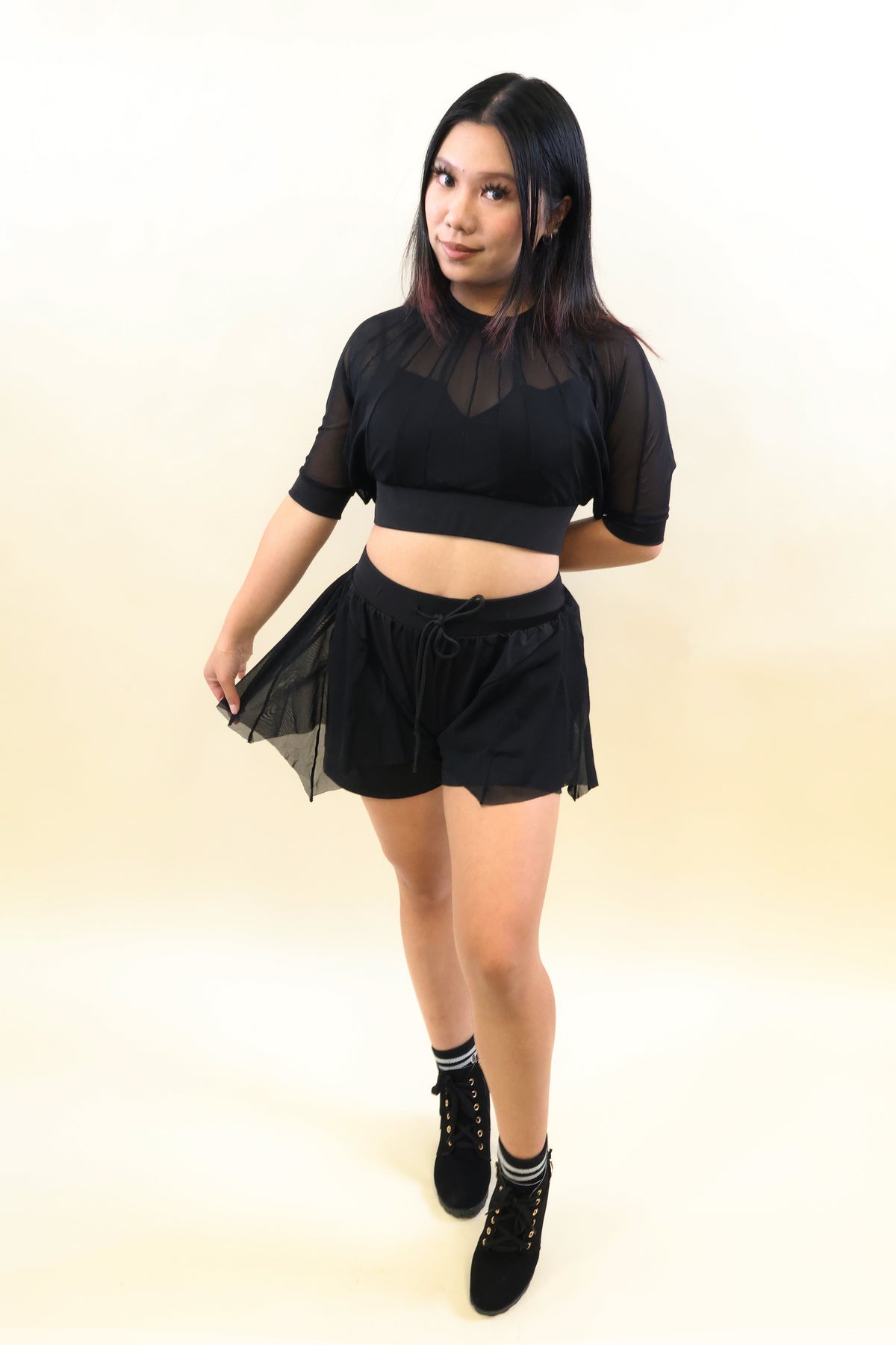 A woman modeling a black two-piece outfit consisting of a sheer mesh short-sleeve crop top and matching black shorts, paired with black lace-up ankle boots, against a plain cream-colored studio background.