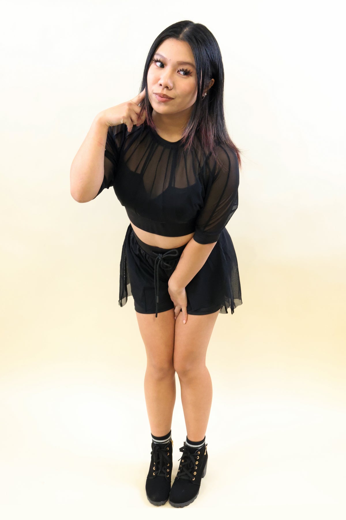 A woman modeling a black sheer mesh short-sleeve crop top and matching black tulle mini skirt, paired with black lace-up ankle boots, against a plain light beige studio background.