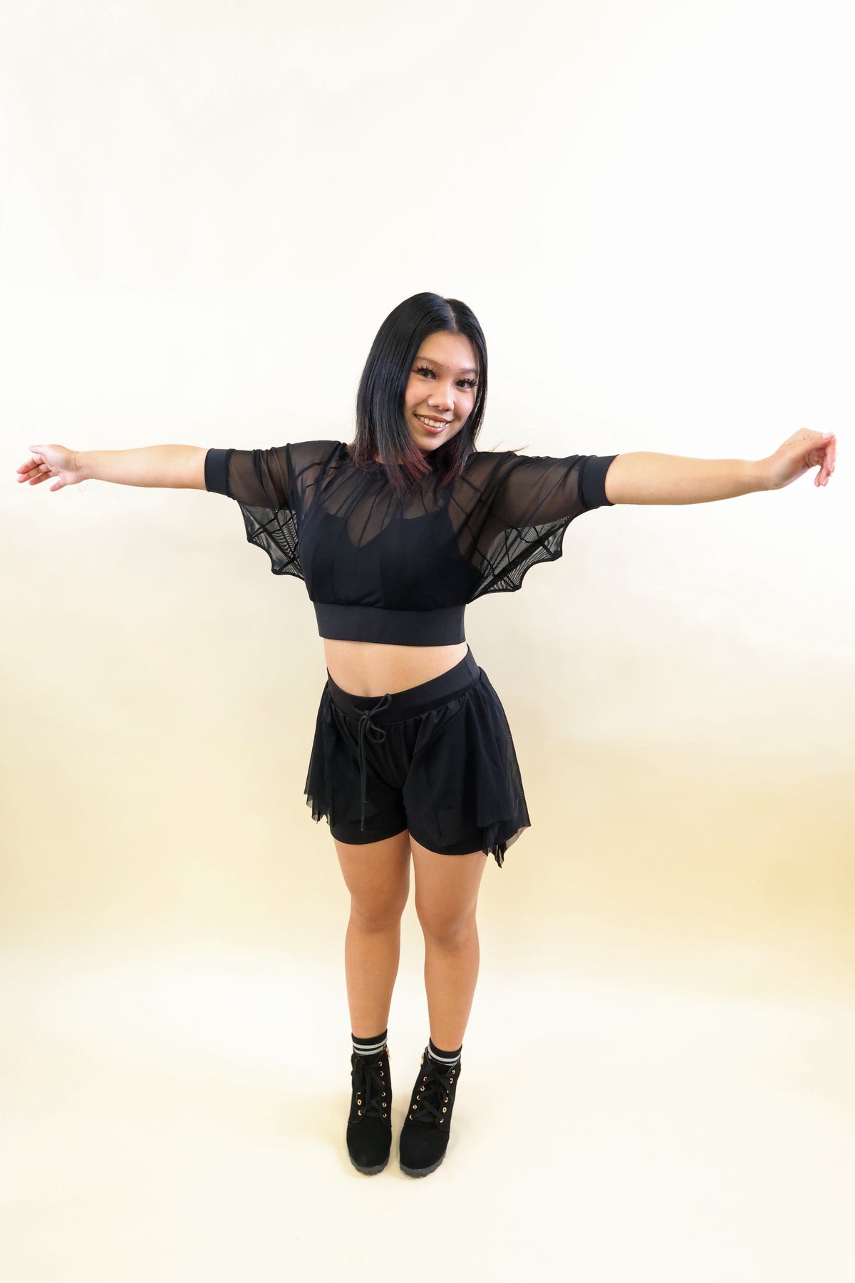 A woman modeling a black sheer mesh crop top with wing-like sleeves and matching black tulle shorts, standing against a plain light beige studio background with her arms outstretched.