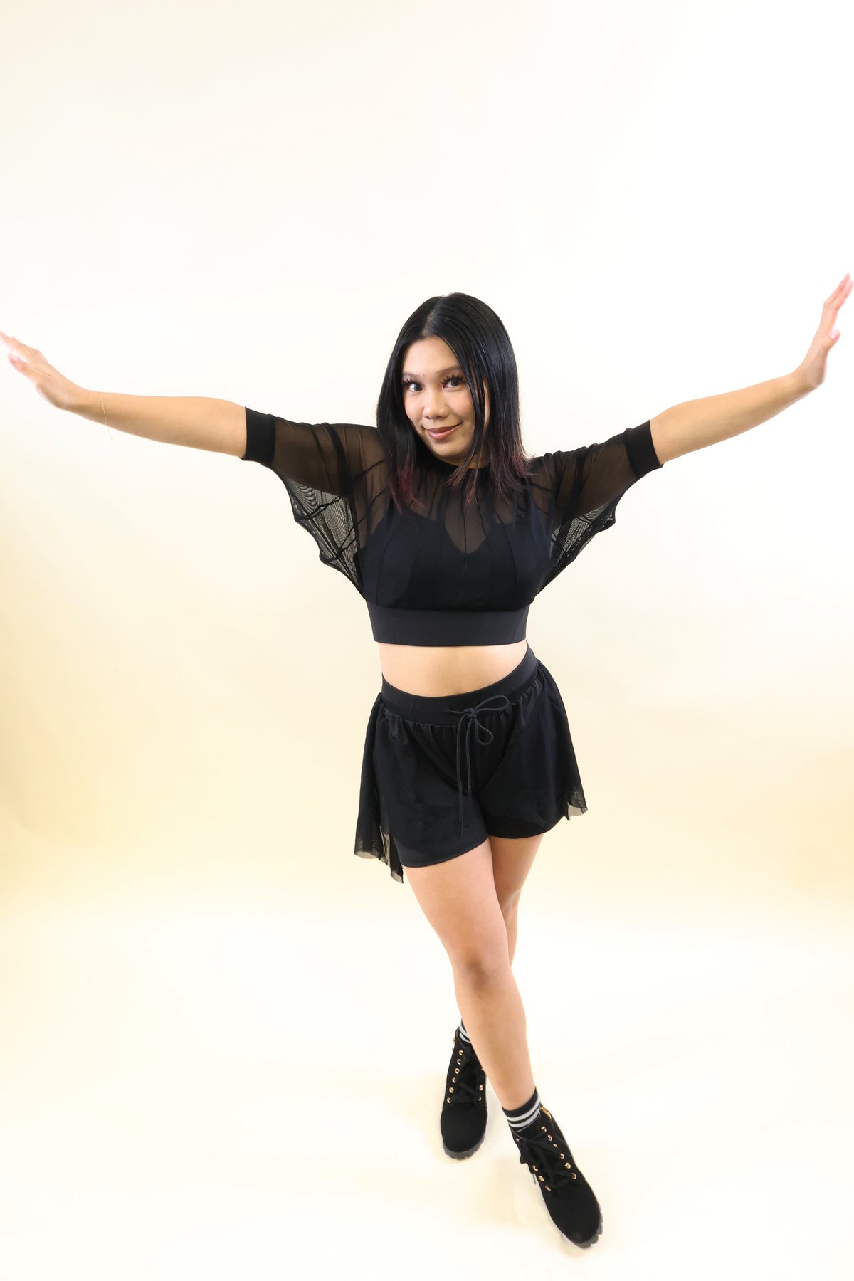A woman modeling a black two-piece dance outfit consisting of a sheer short-sleeve crop top and matching black tulle shorts, posed with her arms outstretched against a plain cream studio background.