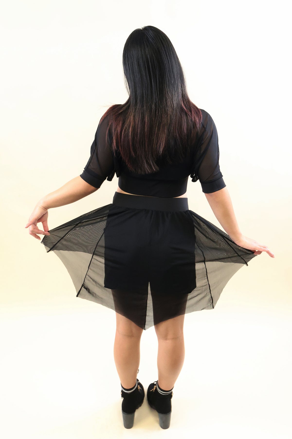 A woman viewed from behind wearing a black sheer mesh short-sleeve crop top and matching black sheer skirt, holding out the sides of the skirt to show its semi-transparent drape. She is wearing black ankle boots against a plain white studio background.