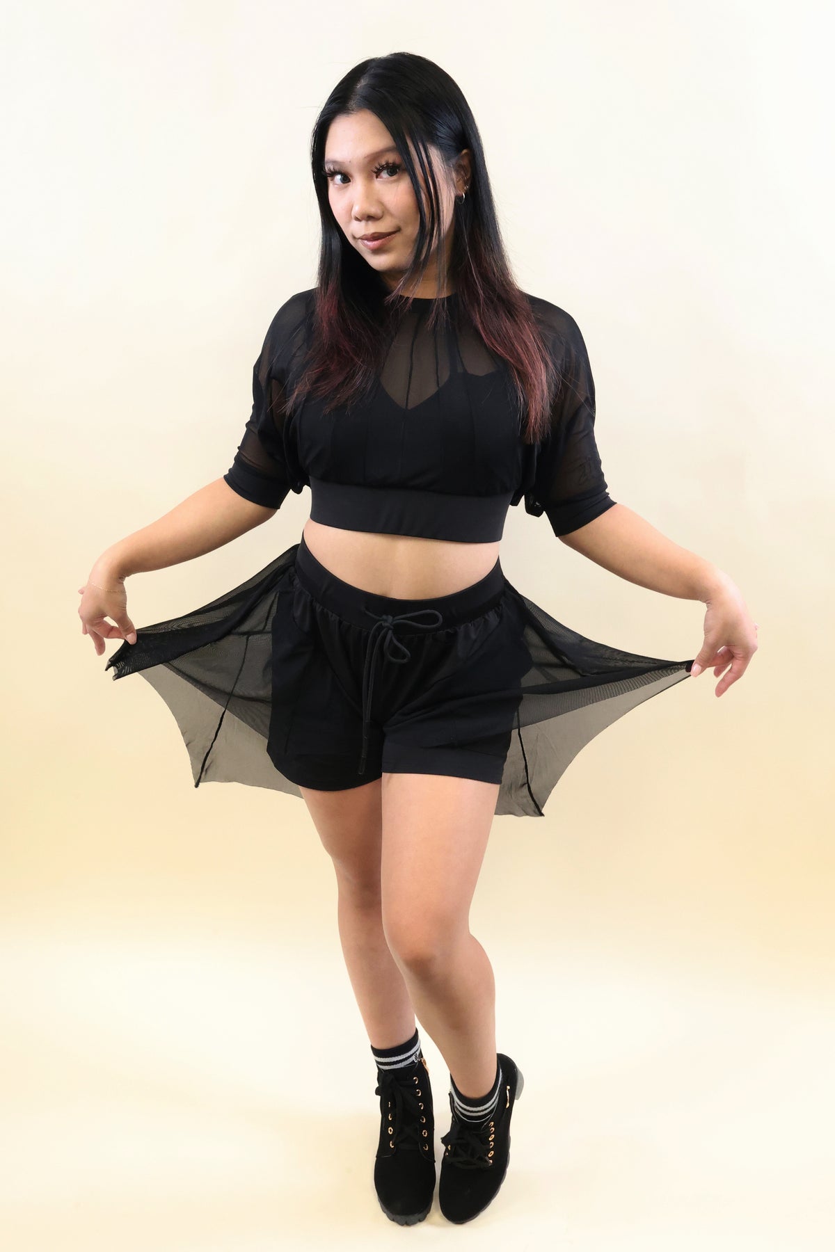 A woman modeling a black sheer mesh two-piece outfit consisting of a short-sleeve crop top and matching drawstring shorts, posing against a light beige studio background.