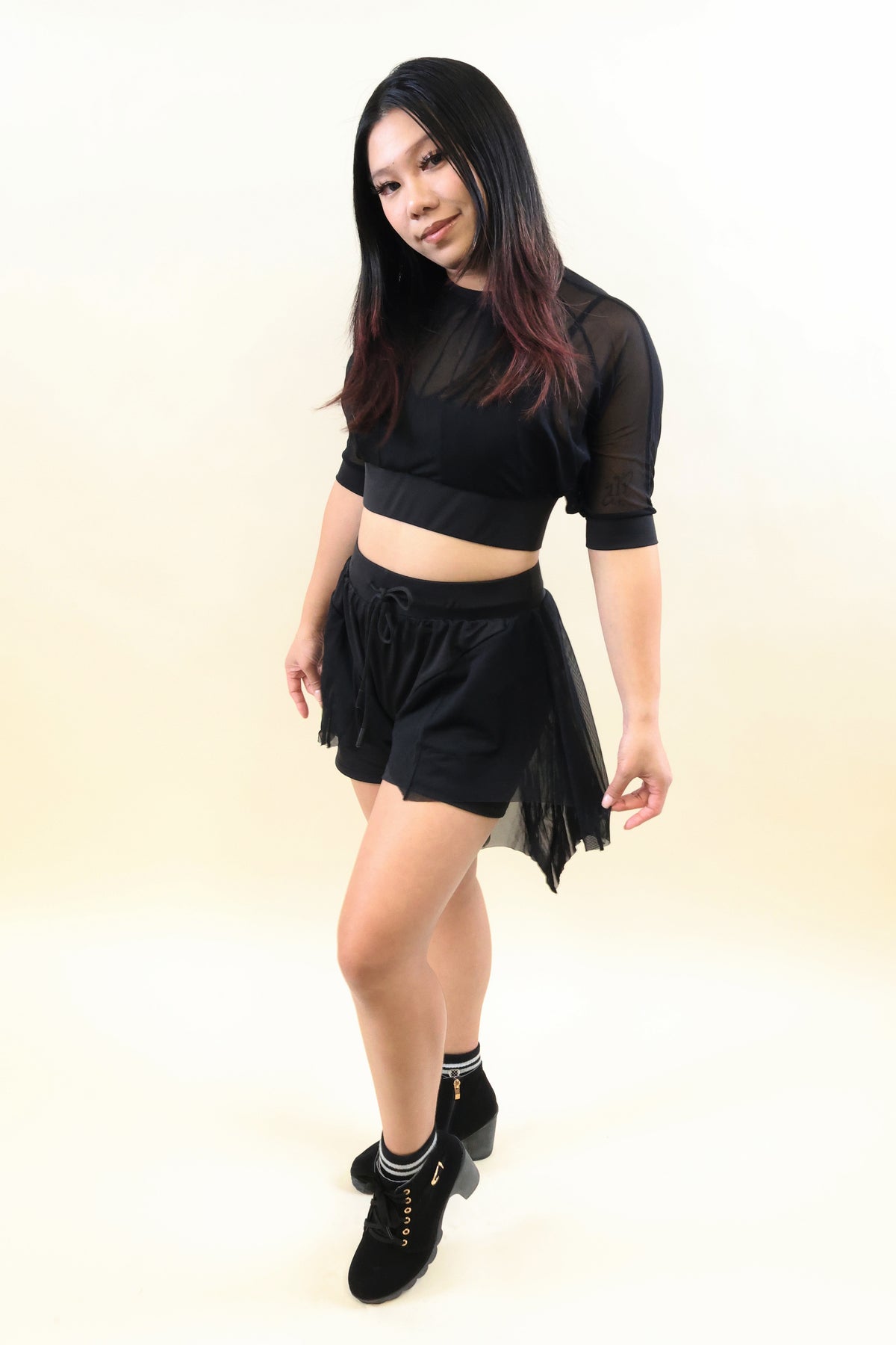 A woman modeling a black two-piece outfit consisting of a sheer short-sleeve crop top and matching black mesh shorts, paired with black socks and black lace-up shoes, against a plain light beige studio background.
